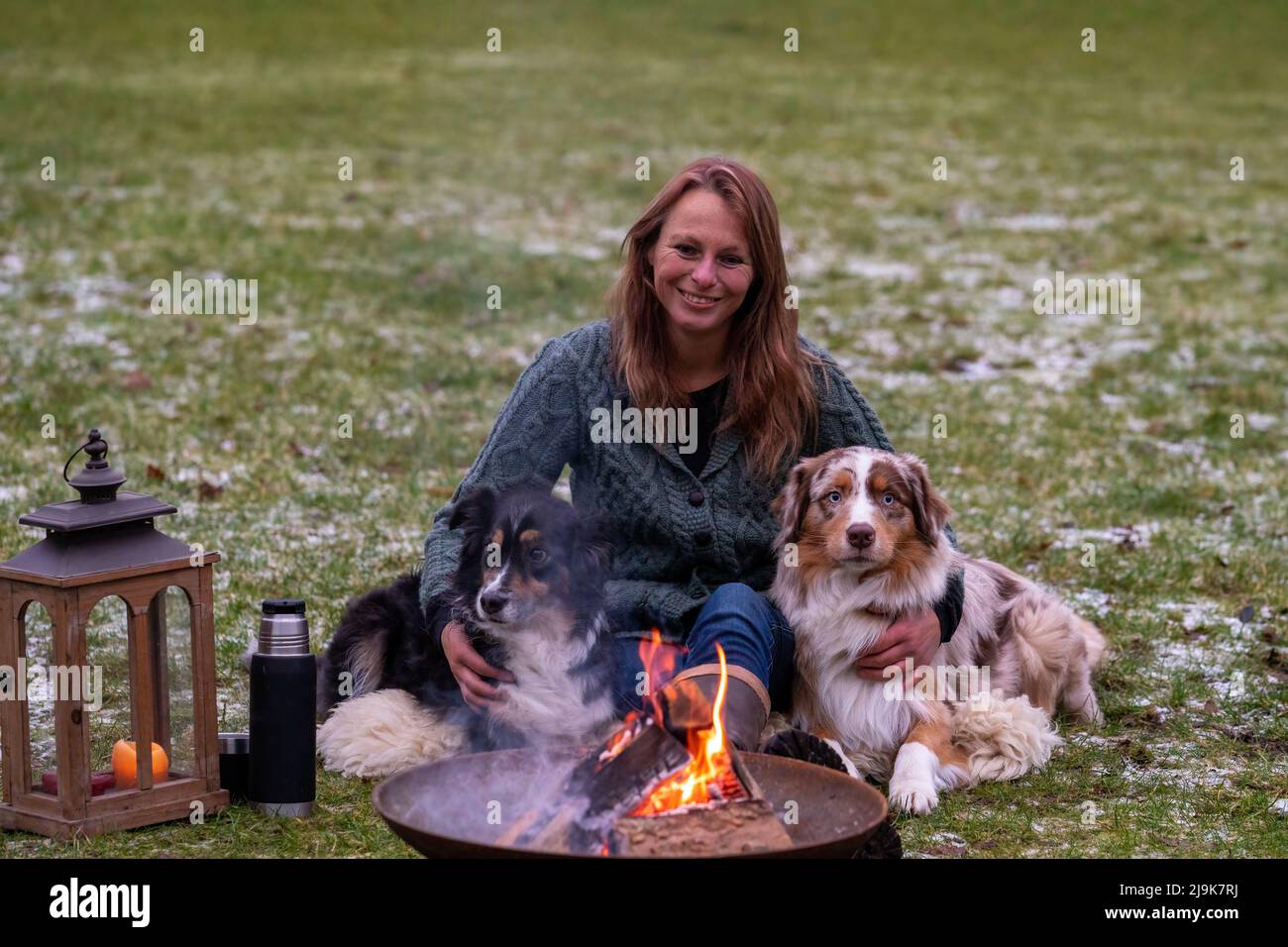 Une jeune femme est assise à l'extérieur dans les bois avec ses deux chiens de berger australiens.Neige sur l'herbe, crépuscule près du feu de camp.Câlins et compagnon Banque D'Images