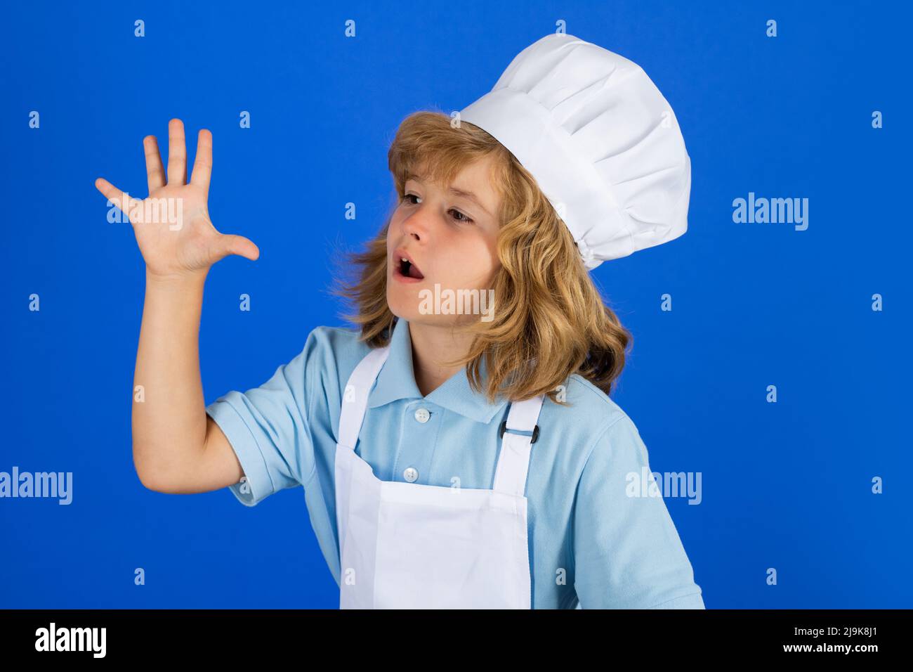 Le chef cuisinier prépare la nourriture dans un décor de studio bleu isolé. La cuisine des enfants. Un garçon de l'adolescence avec un tablier et un chapeau de chef préparant un repas de légumes sains Banque D'Images