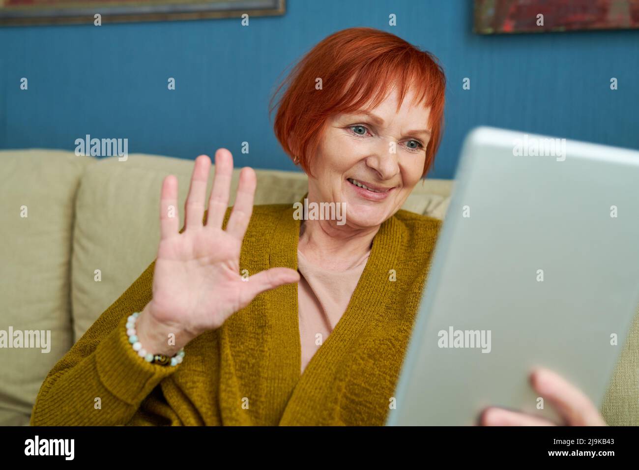 Une femme mûre agitant la main tenant une tablette numérique tout en étant assise sur un canapé à la maison, elle appelle sa famille en ligne Banque D'Images