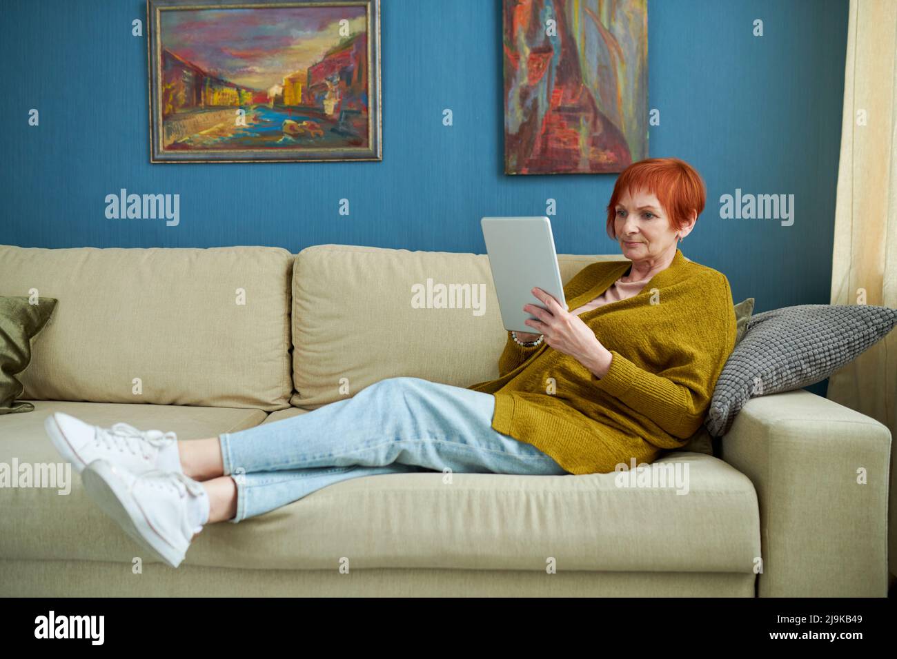 Femme âgée couchée sur un canapé dans le salon et utilisant une tablette numérique, elle communique en ligne ou regarde la vidéo Banque D'Images