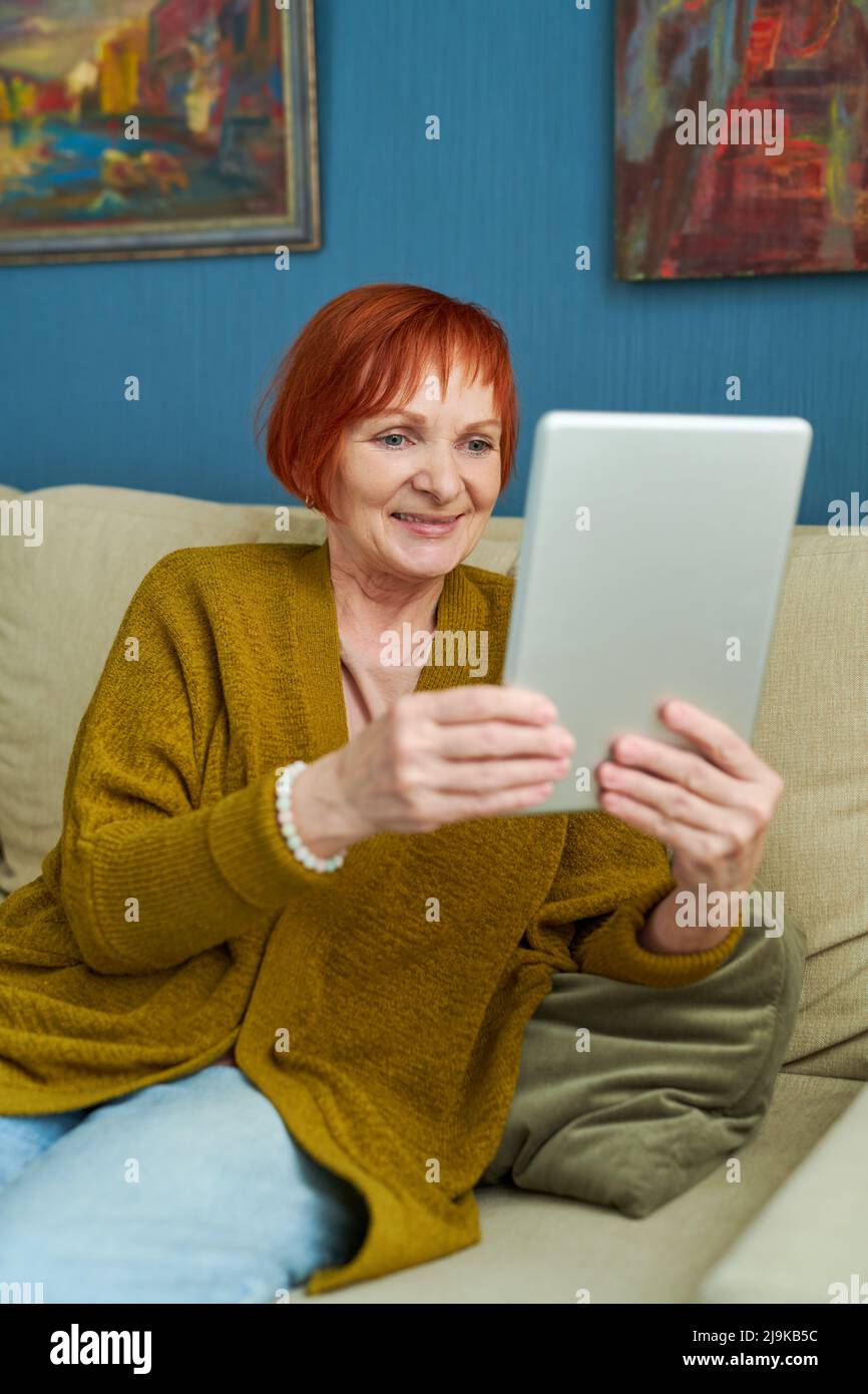 Femme sénior aux cheveux, assise sur un canapé et tenant un Tablet pc devant elle lors d'une conversation en ligne avec sa famille Banque D'Images
