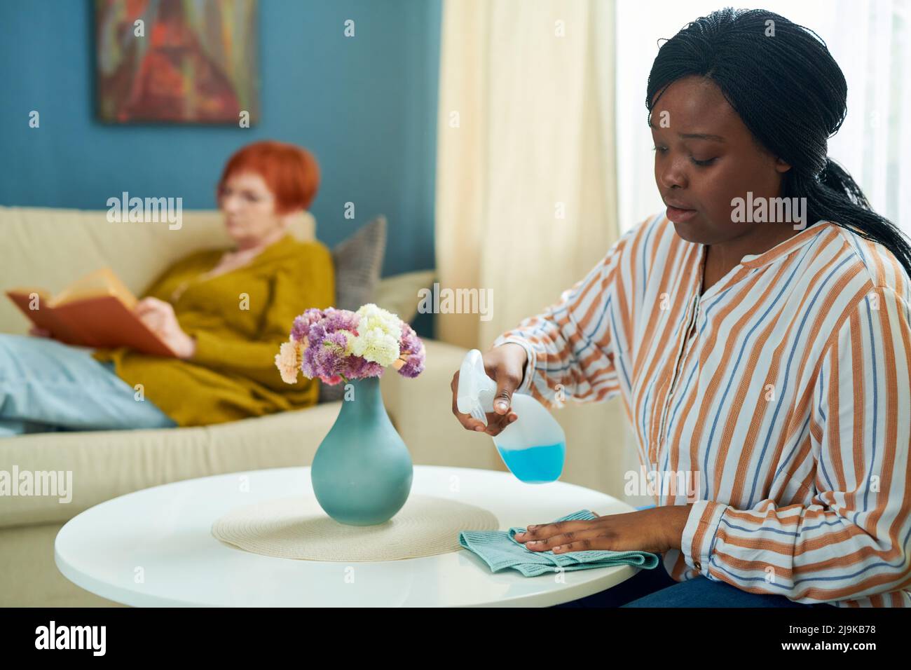 Jeune volontaire africain aidant une femme âgée à la maison, elle essuyant la poussière de la table et faisant des travaux ménagers Banque D'Images