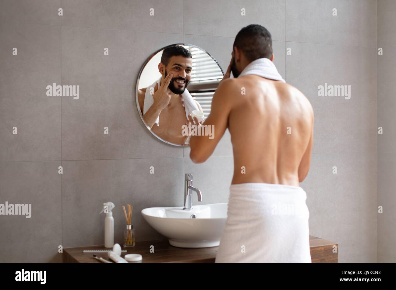Joyeux gars regardant dans le miroir mettant la crème sur le visage Banque D'Images