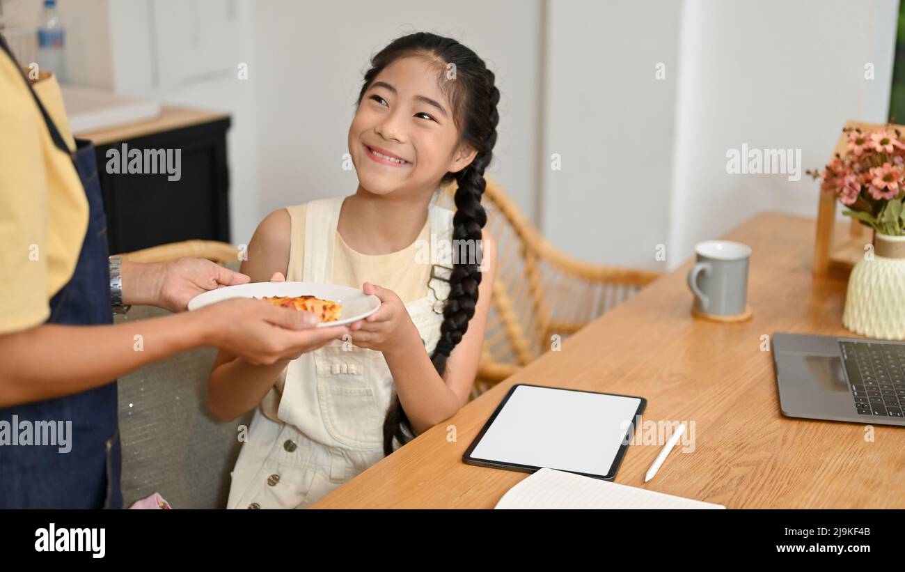 Charmant concept de famille asiatique. Maman donne du pain à sa fille pendant qu'elle étudie en ligne. Une fille souriante s'assoit à la table à manger et reçoit une assiette de brea savoureuse Banque D'Images