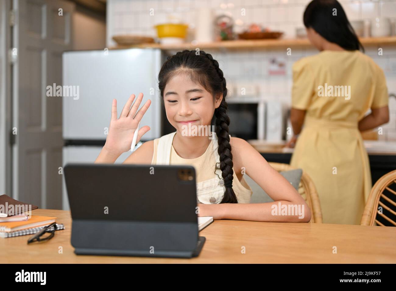 Jolie jeune fille asiatique étudiant sur le cours en ligne via un ordinateur tablette, s'assoit dans la salle à manger tandis que sa mère cuisine dans la cuisine derrière. Banque D'Images