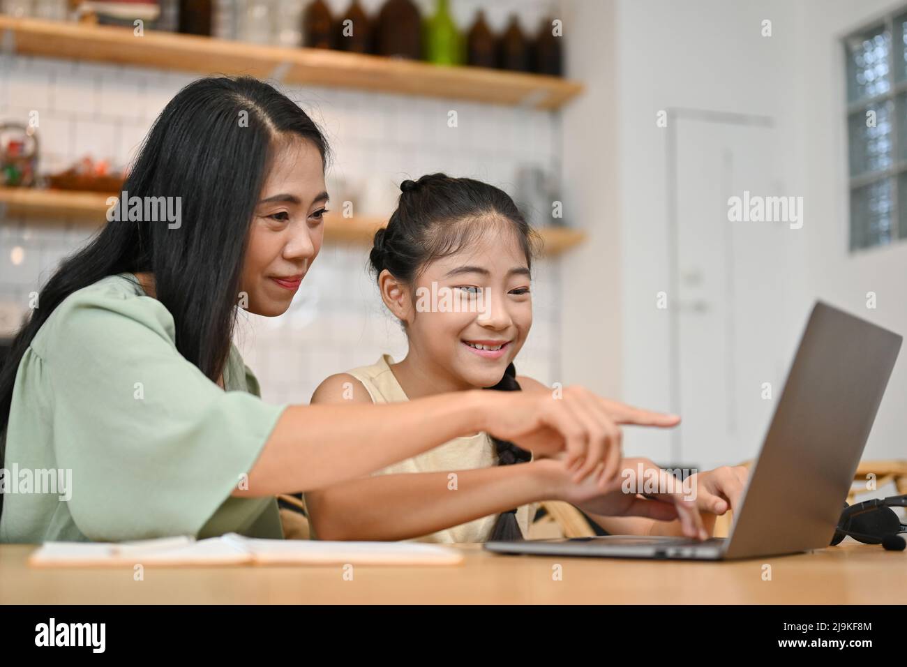 Maman asiatique gaie regardant l'écran d'un ordinateur portable, aidant sa fille à faire ses devoirs en ligne, expliquant la question et disant la réponse. Banque D'Images