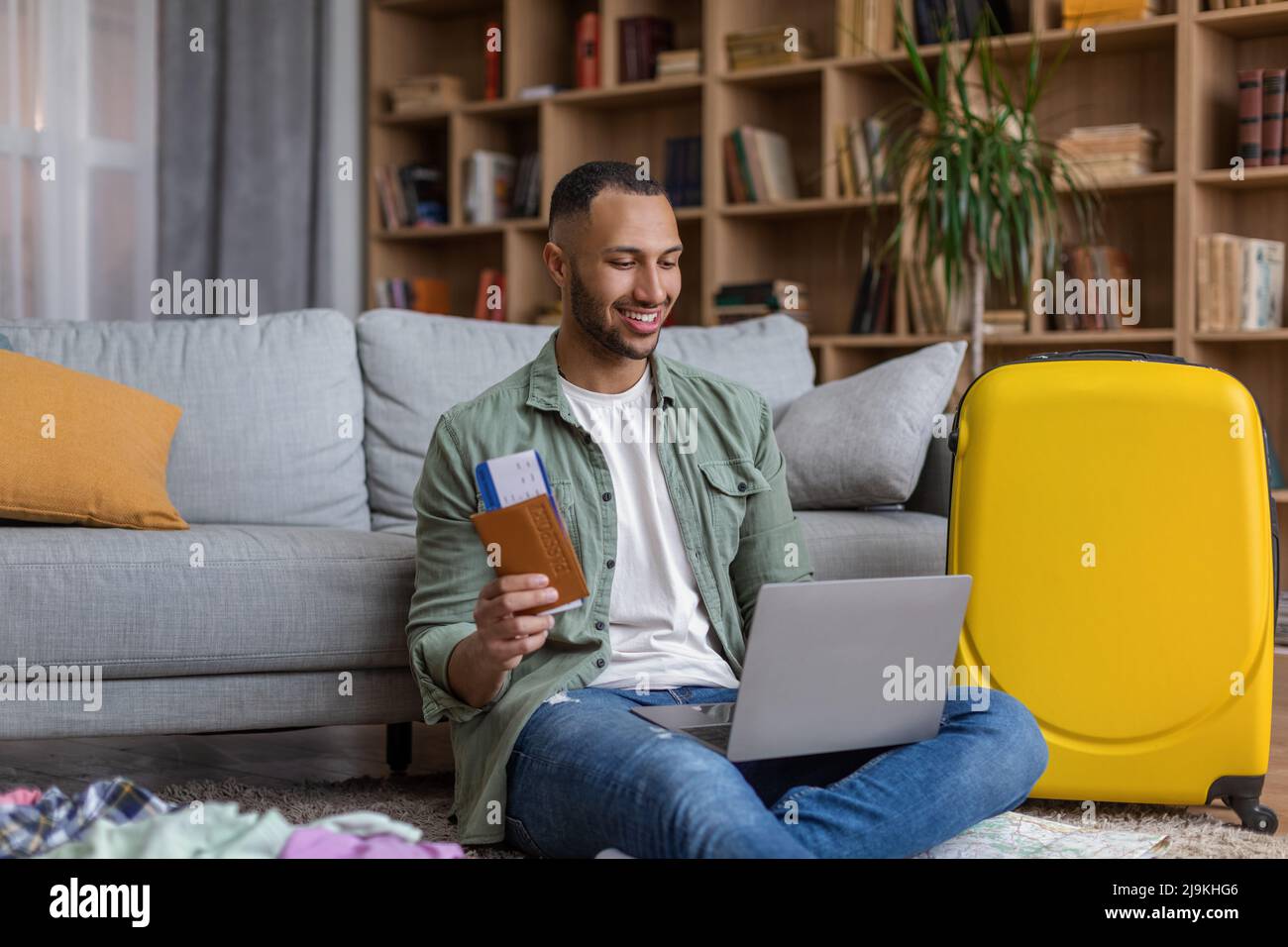 Concept de service en ligne de l'agence de voyages. Un homme noir positif tenant un billet et un passeport, utilisant un ordinateur portable pour la réservation d'hôtel Banque D'Images