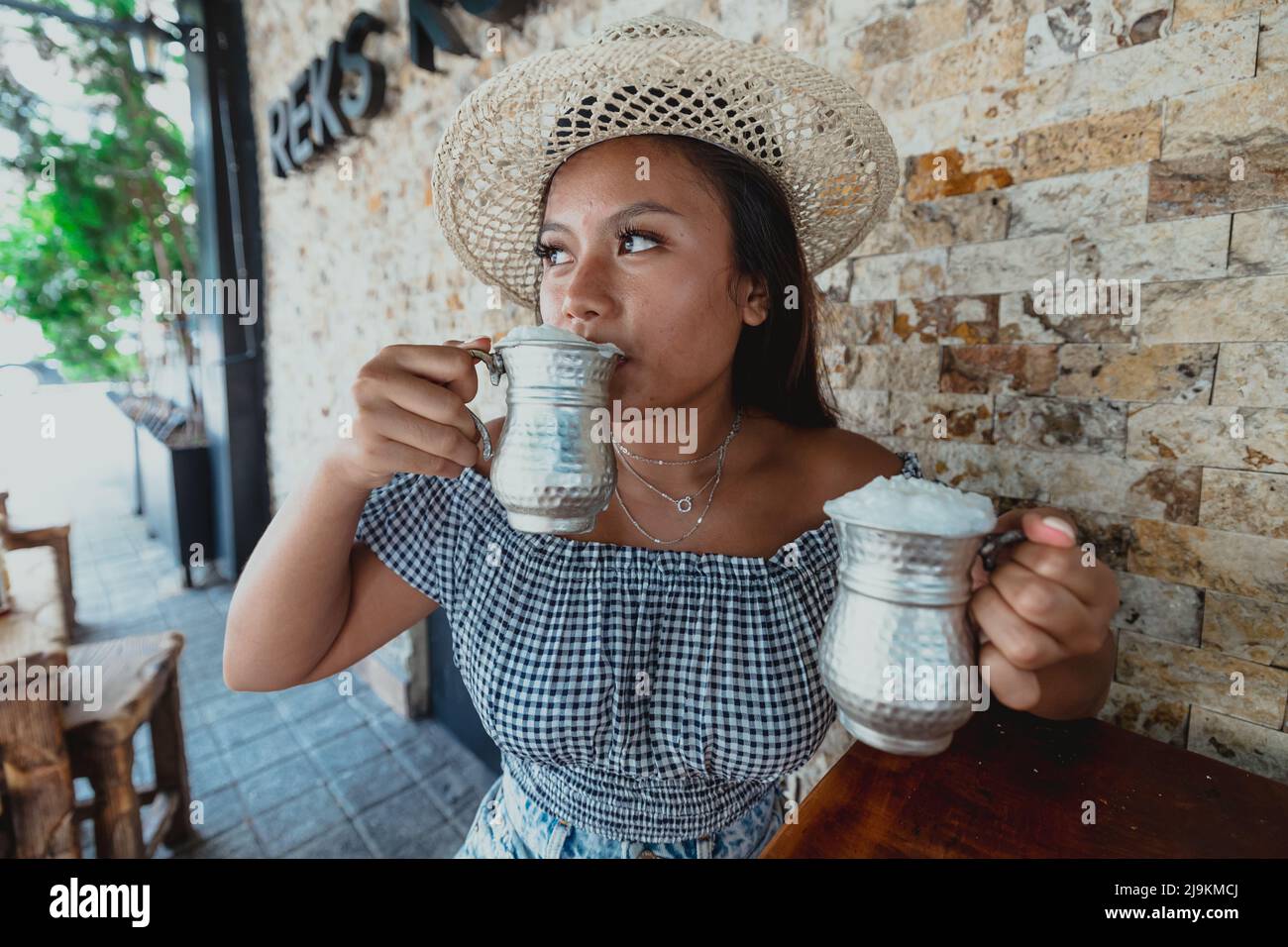 une jeune fille indonésienne buvant de l'ayran dans une tasse d'argent portant un chapeau pendant l'été Banque D'Images