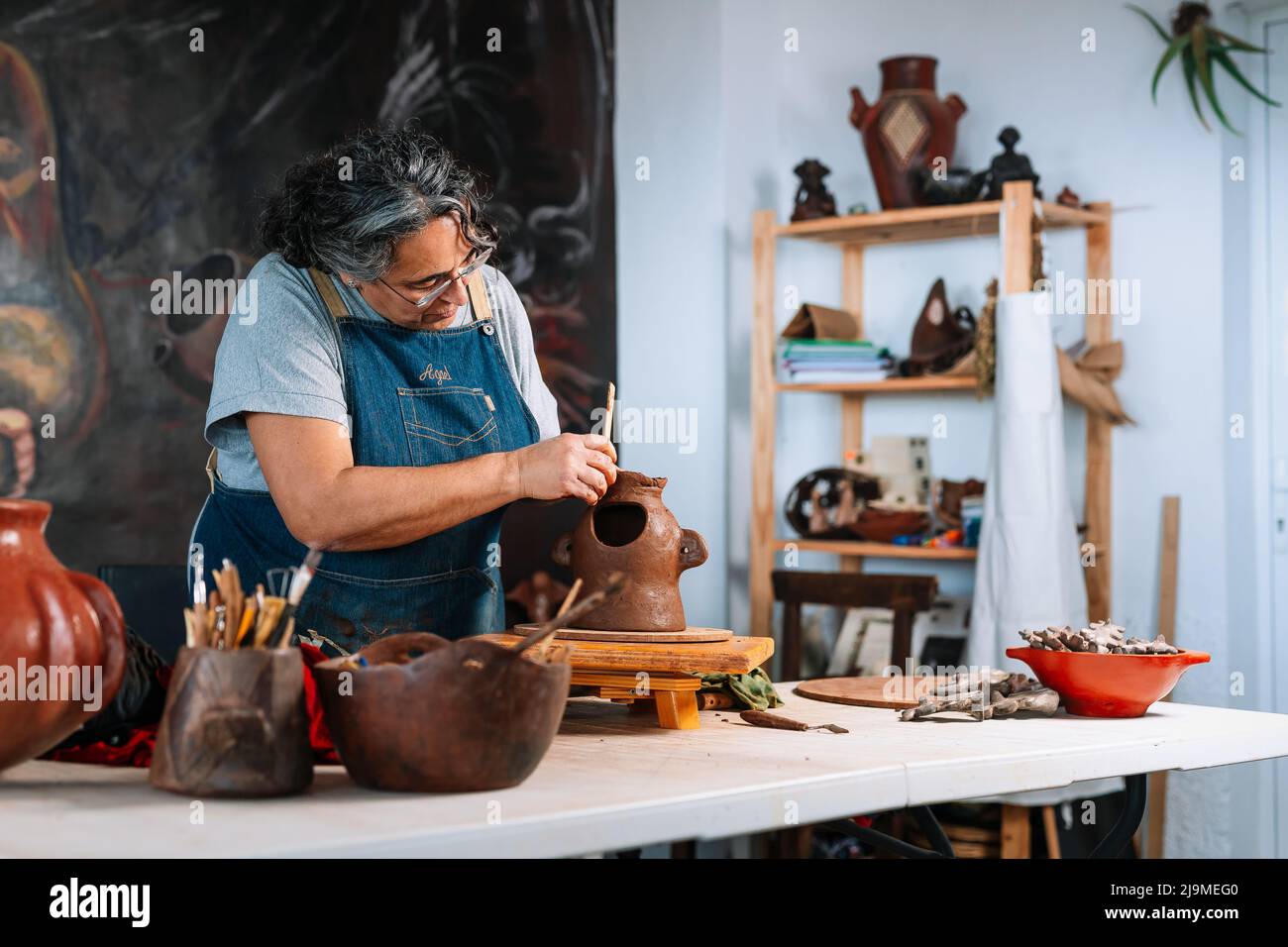 Vue latérale d'une femme mûre et centrée sur l'artisan Ceramist Woman en tablier façonnant la pièce de pot d'argile avec l'outil dans la table d'atelier Banque D'Images