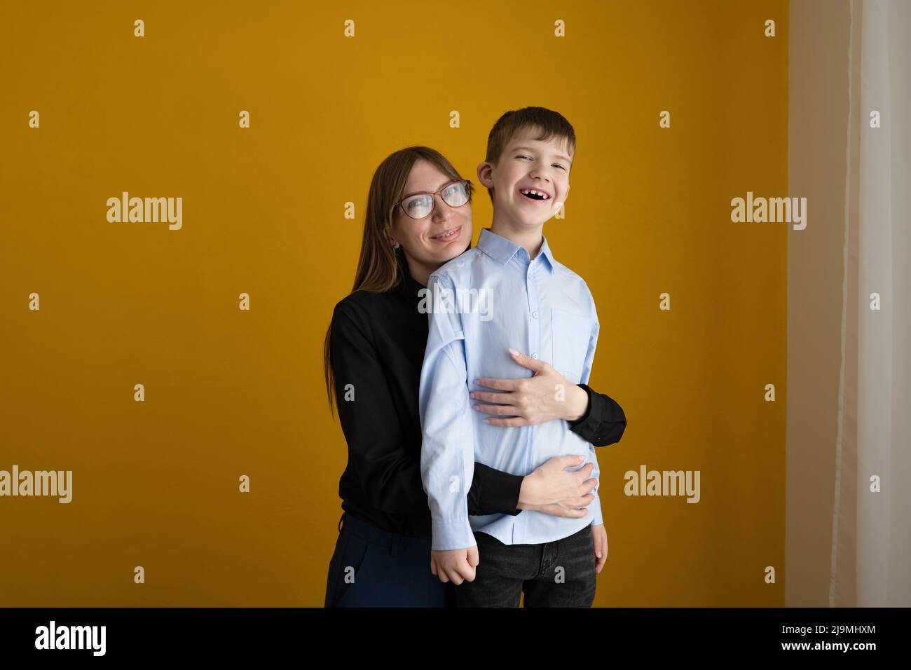 Maman adulte dans des vêtements et des lunettes élégants et décontractés embrassant enfant optimiste de derrière tout en se tenant contre le mur jaune à la maison Banque D'Images