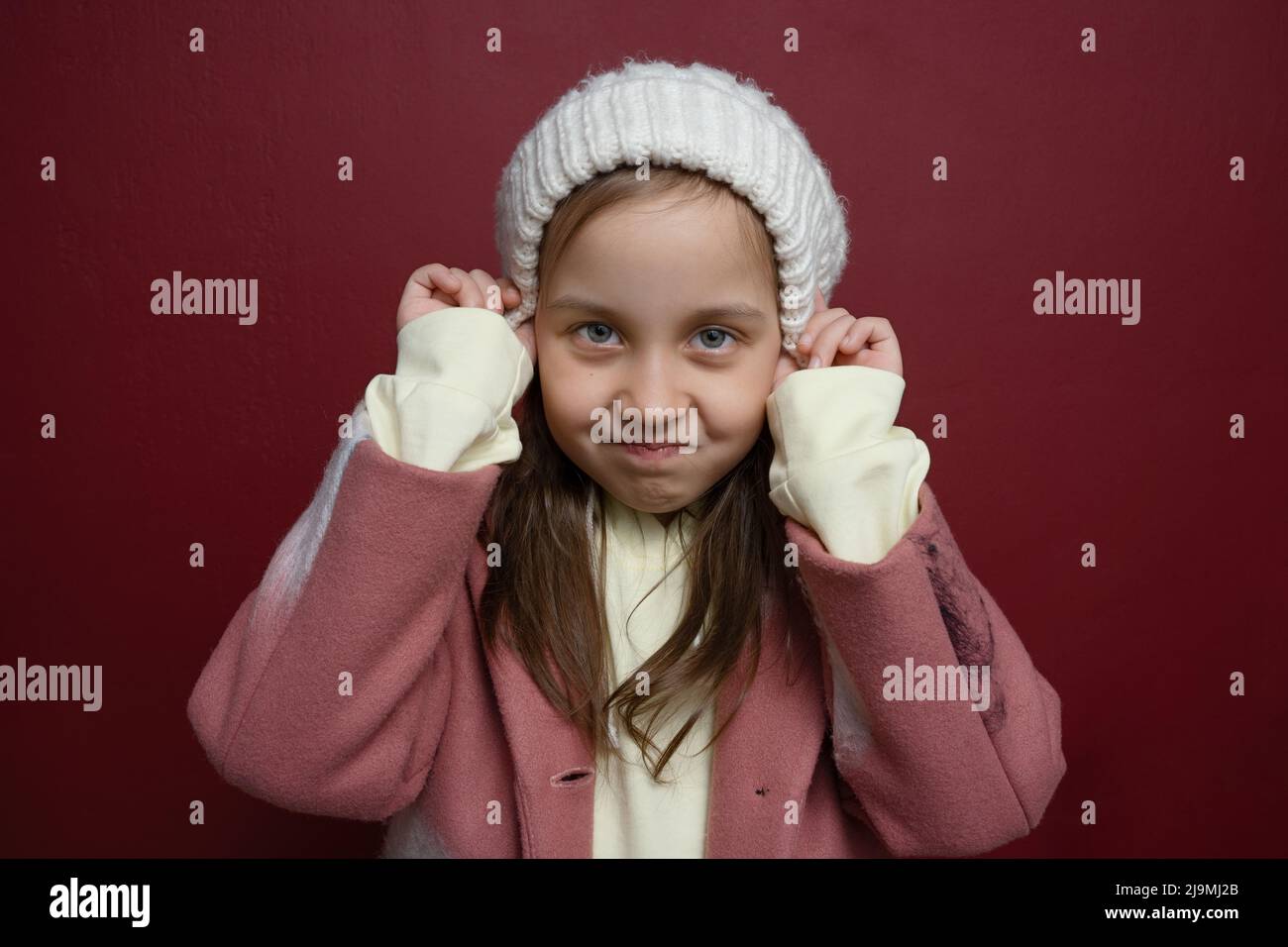 Une petite fille capricieuse avec des cheveux longs en vêtements d'extérieur tendance mettant sur un bonnet tricoté chaud et regardant l'appareil photo avec insatisfaction contre le dos bordeaux Banque D'Images