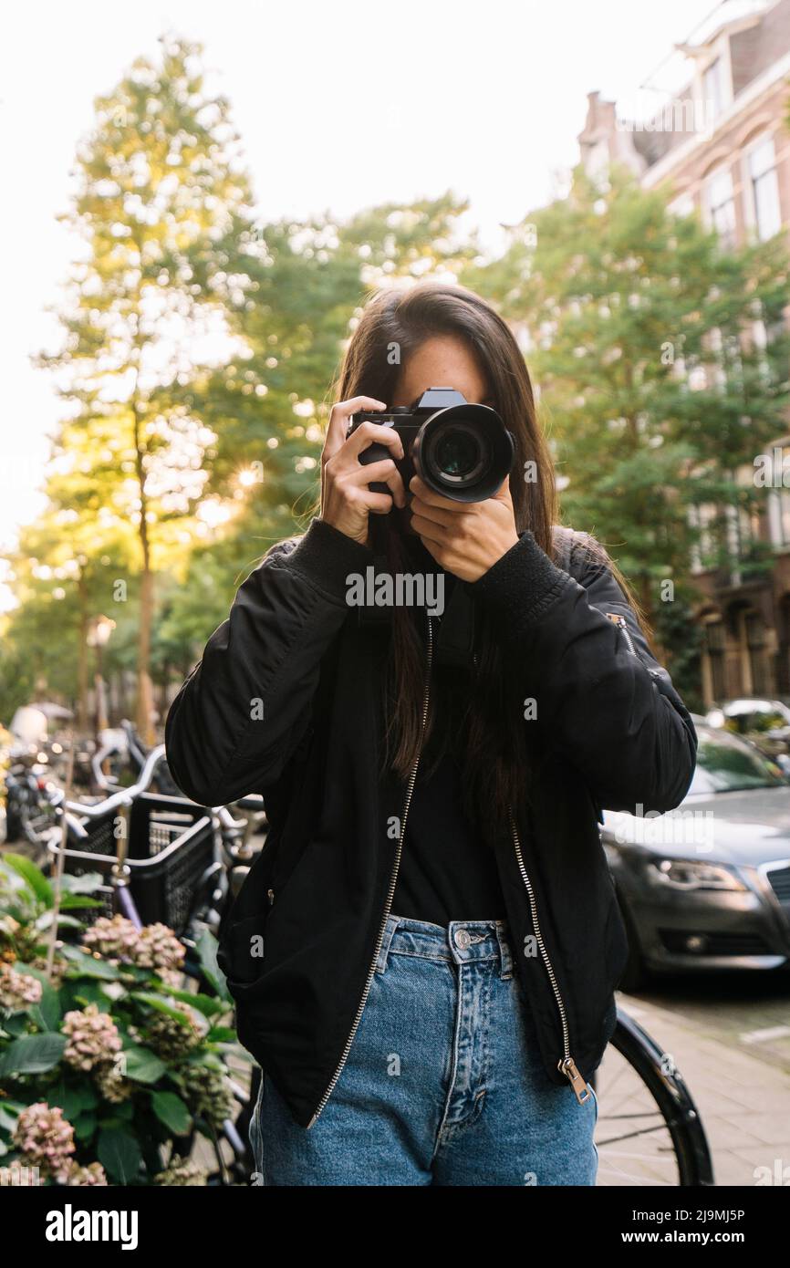 Jeune touriste femelle méconnaissable avec de longs cheveux foncés dans une veste élégante prenant des photos sur un appareil photo tout en se tenant dans la rue près de la dur à vélo garée Banque D'Images