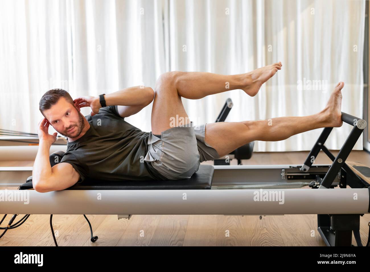 Vue latérale d'un athlète pieds nus allongé sur un reformeur pilates et effectuant un exercice d'abs pendant un entraînement physique. Pilates MAN concept. Banque D'Images