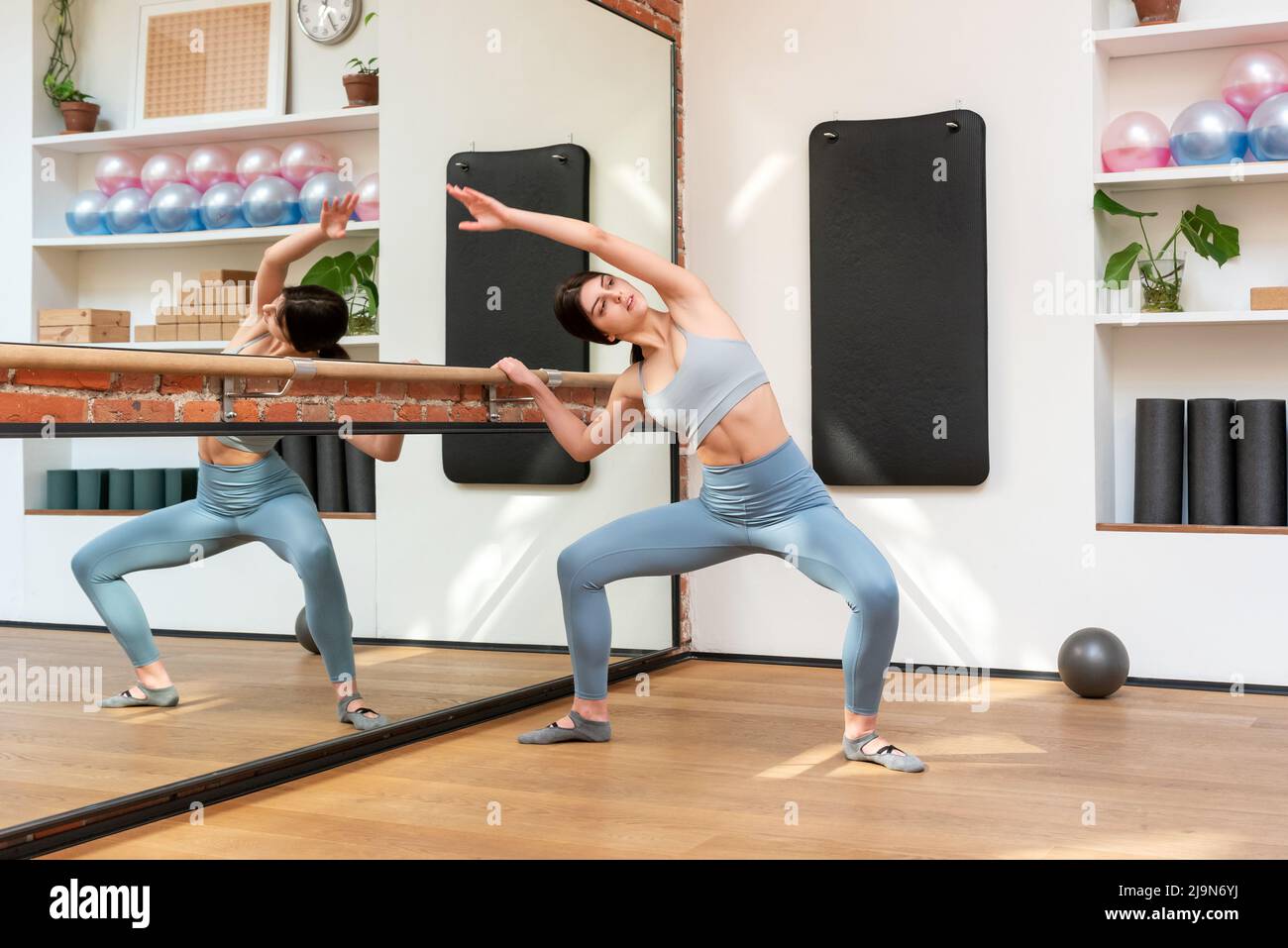 Danseuse de ballet féminine pleine forme dans les vêtements de sport en train de jouer au squat en faisant de l'exercice dans un studio spacieux avec barre et miroir Banque D'Images