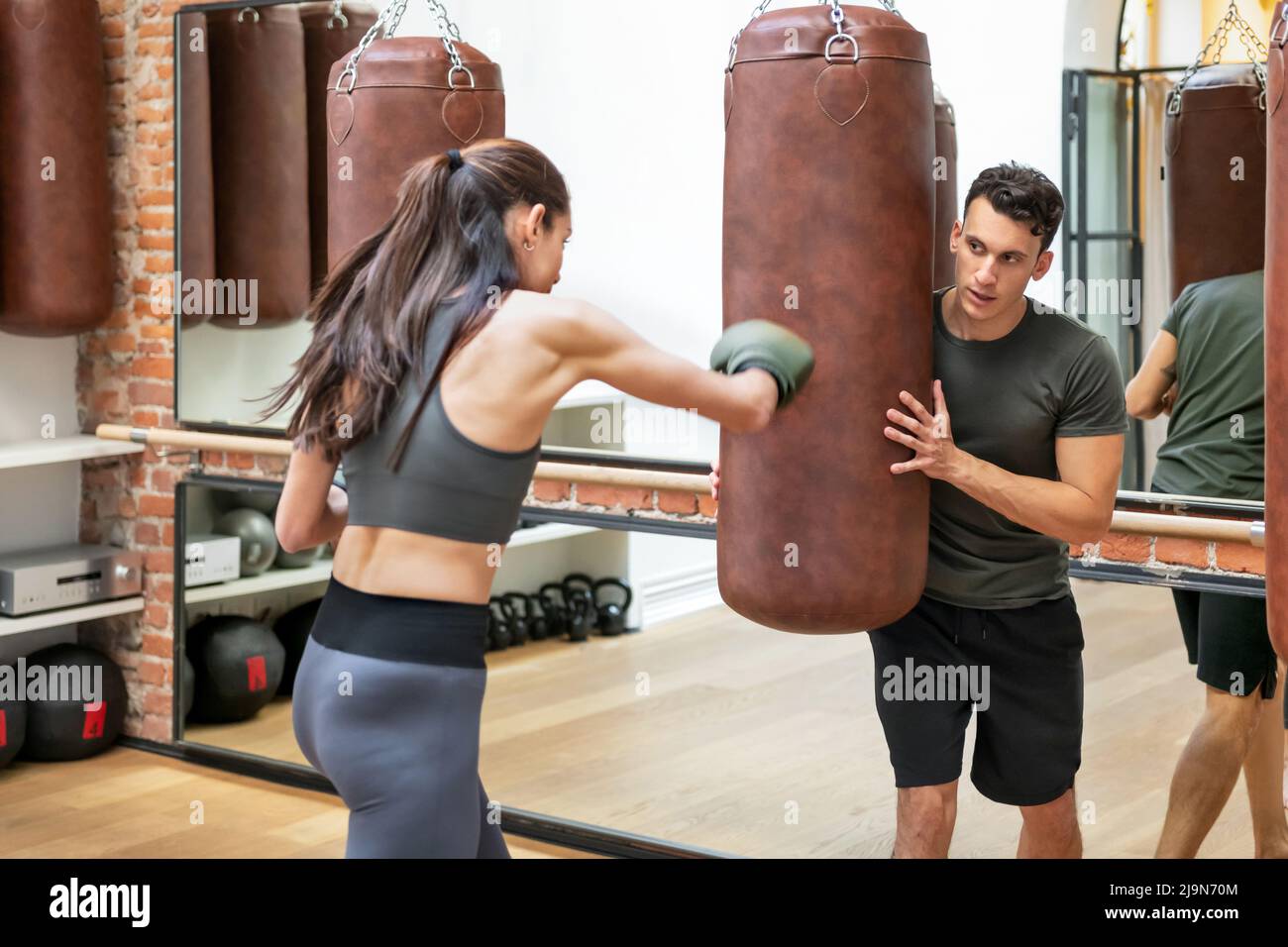 Une femme sportive en mitaines de boxe fait un sac de poinçonnage tout en faisant de l'exercice avec l'aide d'un entraîneur personnel masculin dans un club de fitness moderne Banque D'Images