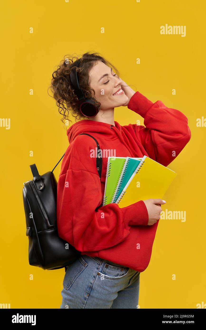 Vue latérale d'une étudiante écoutant de la musique dans des écouteurs. Fille debout avec sac à dos, tenant des livres, regardant vers le haut avec les yeux fermés, souriant. Isolé sur fond jaune. Banque D'Images Vue latérale d'une étudiante écoutant de la musique dans des écouteurs. Fille debout avec sac à dos, tenant des livres, regardant vers le haut avec les yeux fermés, souriant. Isolé sur fond jaune. Banque D'Images