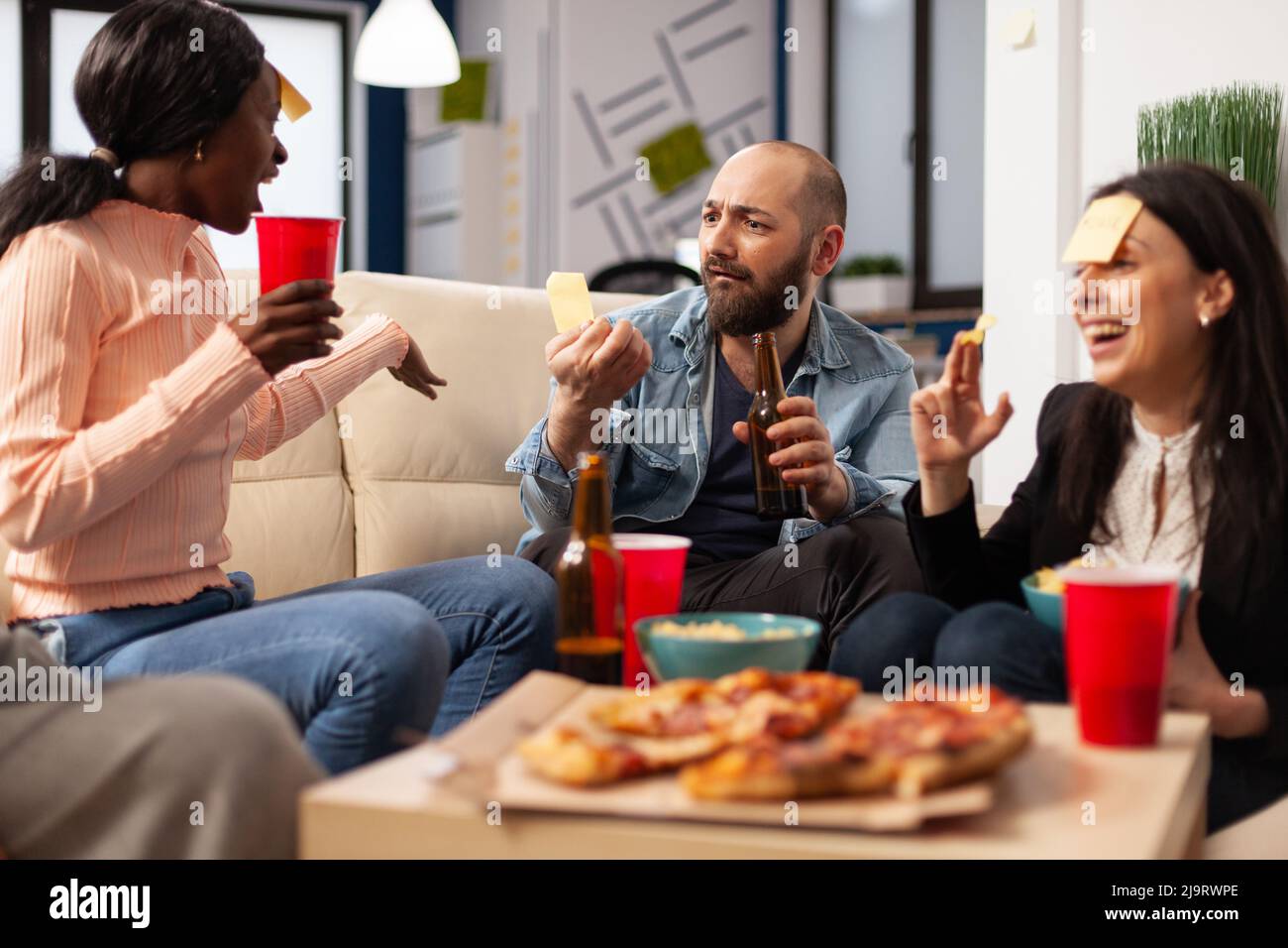 Les collègues de bureau jouant à Guess Who charades jouent à la fête avec des boissons alcoolisées et des en-cas. Des gens joyeux qui utilisent des notes adhésives pour s'amuser et faire des activités de loisirs après le travail. Banque D'Images