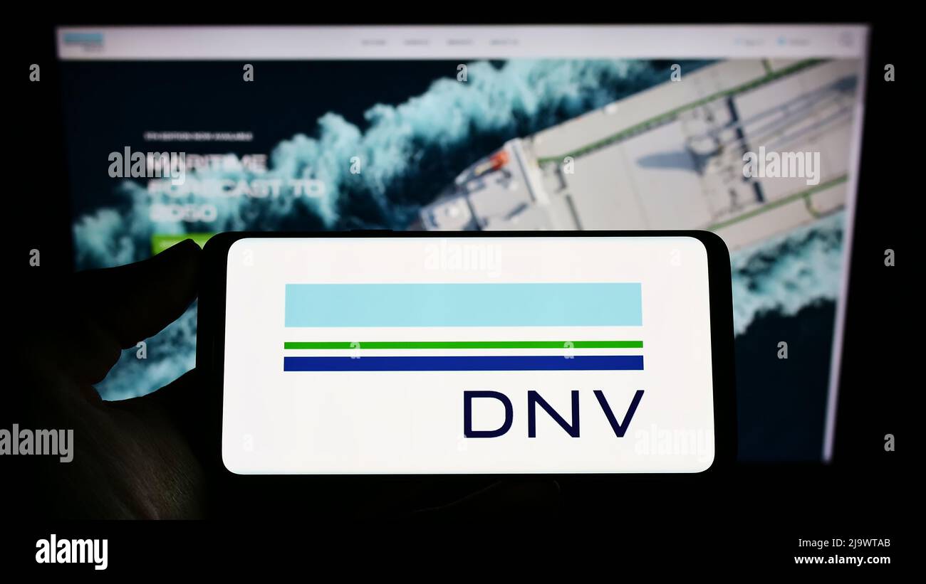 Personne tenant un téléphone mobile avec le logo de la société norvégienne de services DNV COMME sur l'écran en face de la page Web d'affaires. Mise au point sur l'affichage du téléphone. Banque D'Images