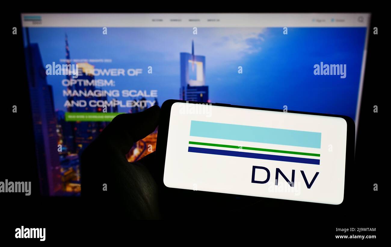 Personne tenant un téléphone portable avec le logo de la société norvégienne de services DNV COMME à l'écran en face de la page Web d'affaires. Mise au point sur l'affichage du téléphone. Banque D'Images