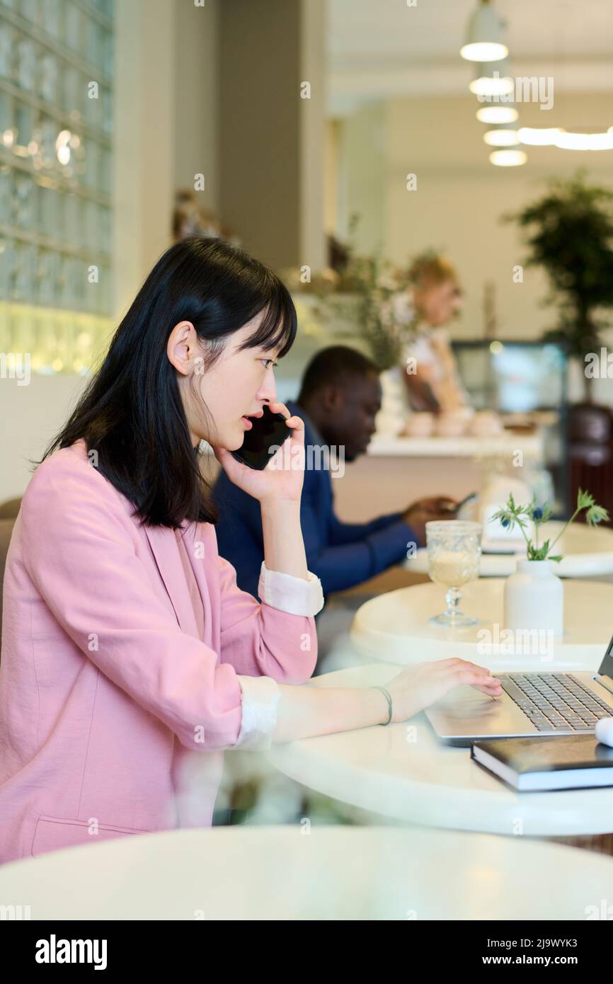 Une jeune femme asiatique parle sur un téléphone portable pendant son travail sur un ordinateur portable à table dans un café Banque D'Images