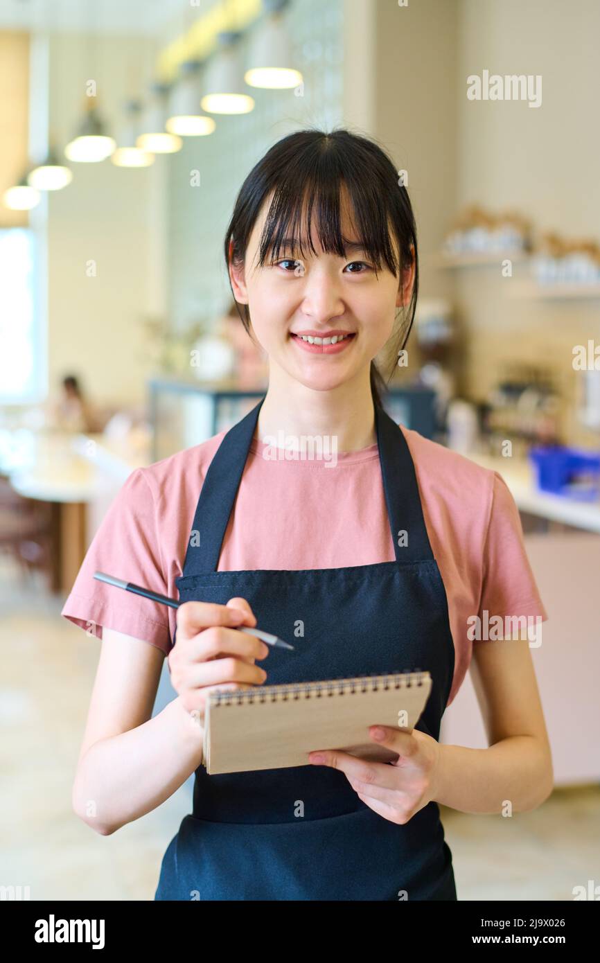 Portrait de la jeune serveuse asiatique en tablier souriant à l'appareil photo tout en prenant la commande au restaurant Banque D'Images