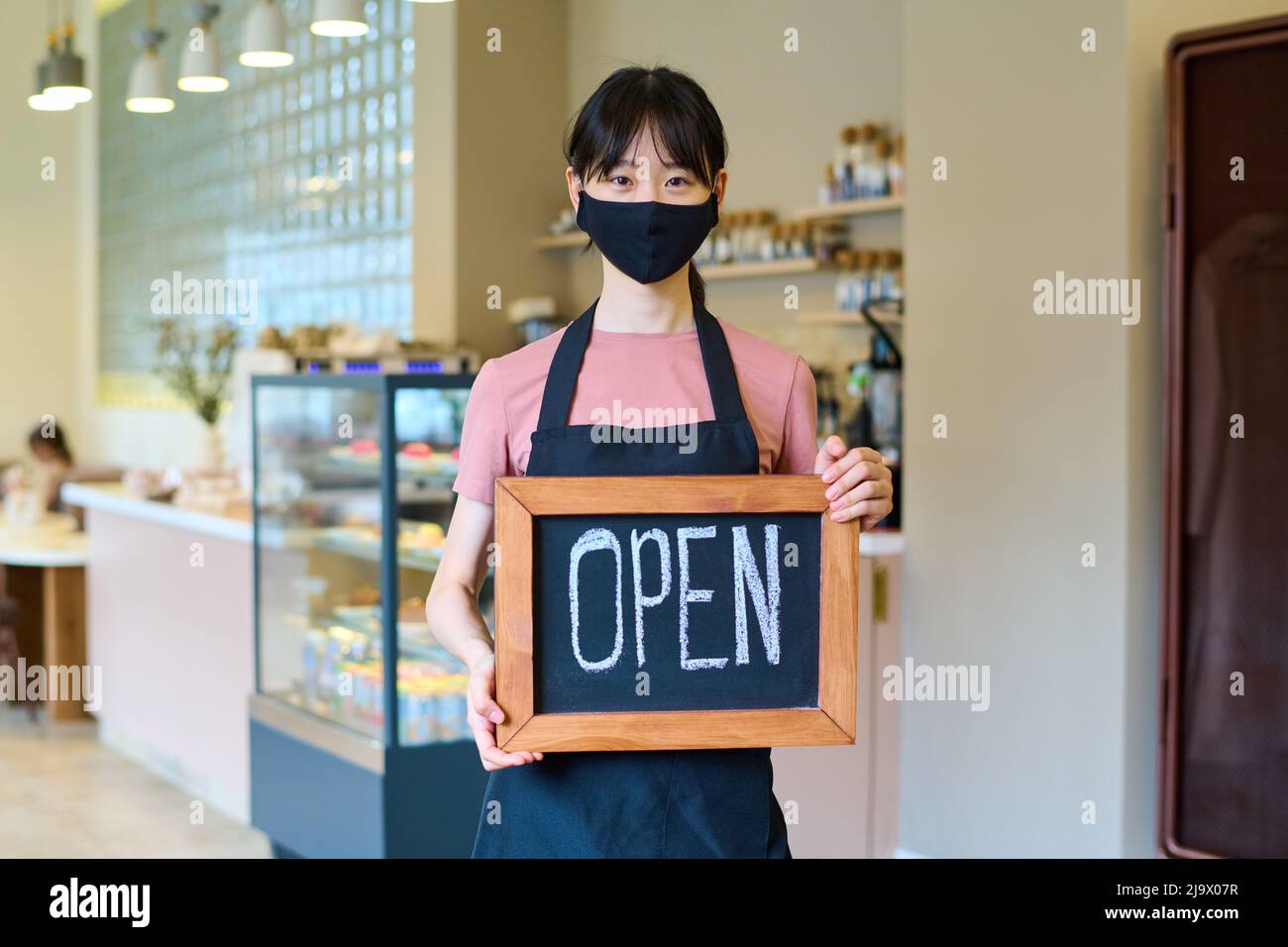 Serveuse asiatique dans un masque facial debout au café portant une affiche ouverte pendant une pandémie de coronavirus Banque D'Images