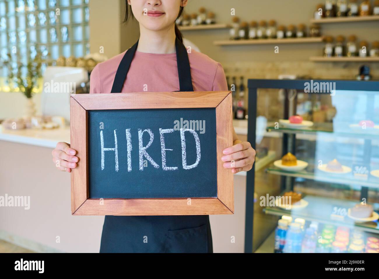 Gros plan de la jeune femme en tablier tenant la planche avec l'enseigne embauchée debout dans le café, elle obtient un nouvel emploi en tant que serveuse Banque D'Images