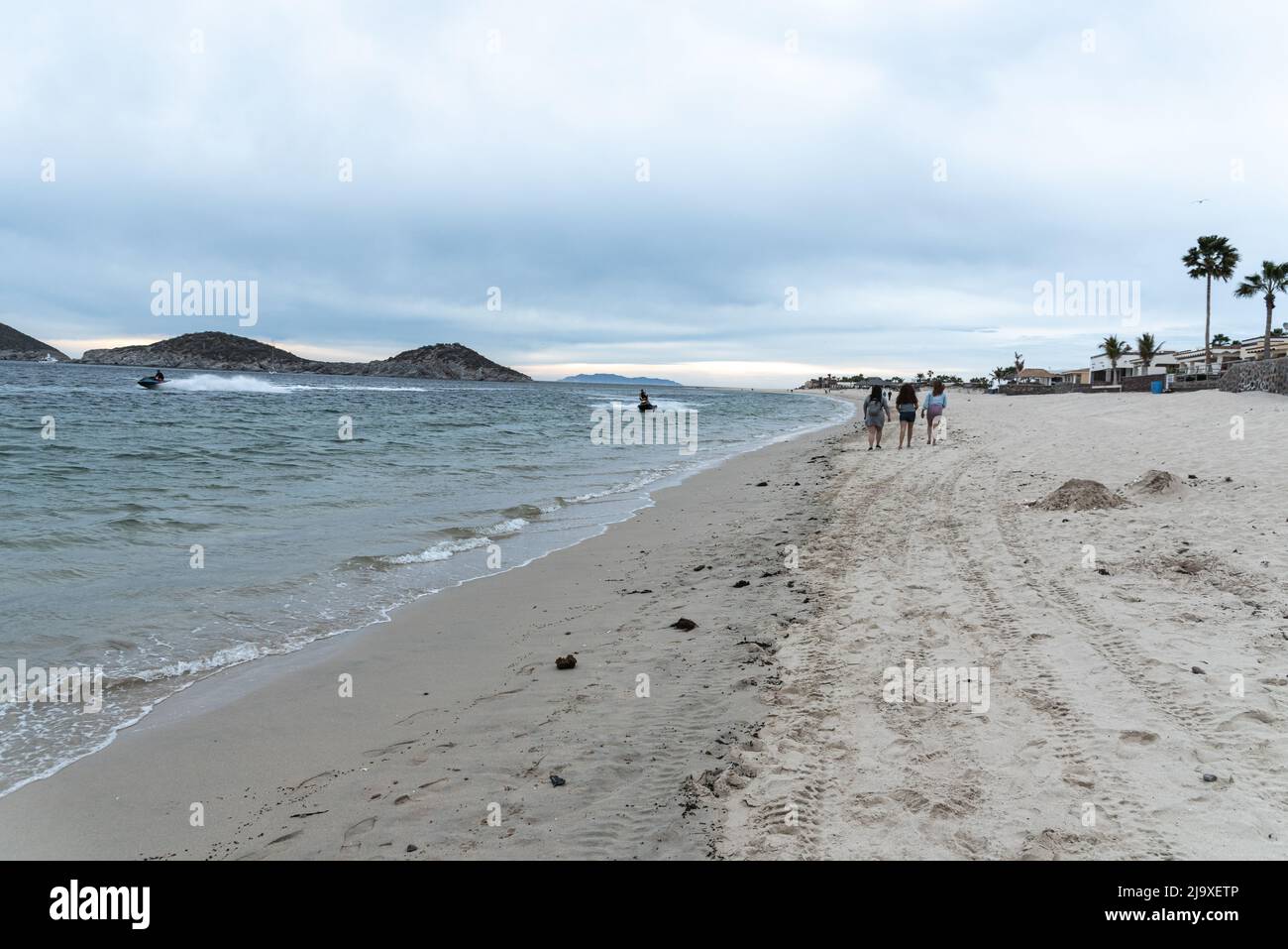 Trois jeunes femmes marchent sur la plage d'Algodones, tandis que deux motomarines se promeulent dans les mers difficiles sous un ciel nuageux à San Carlos, Sonora, Mexique. Banque D'Images