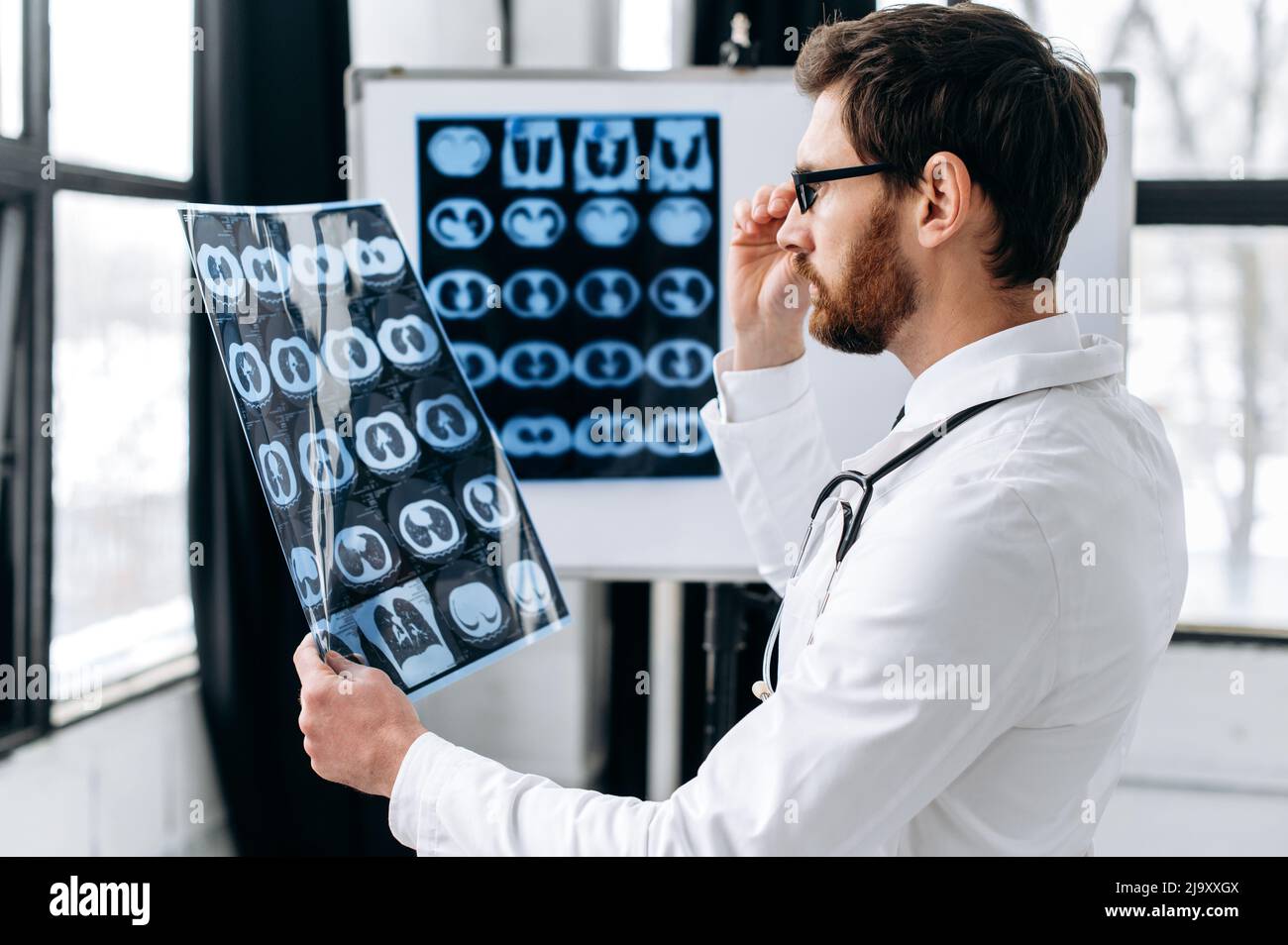 Médecin expérimenté sérieux regardant la radiographie, thérapeute mâle caucasien en uniforme médical blanc et des lunettes concentré de rayons X du patient d'étude, se tient dans le bureau médical. Concept de soins de santé Banque D'Images