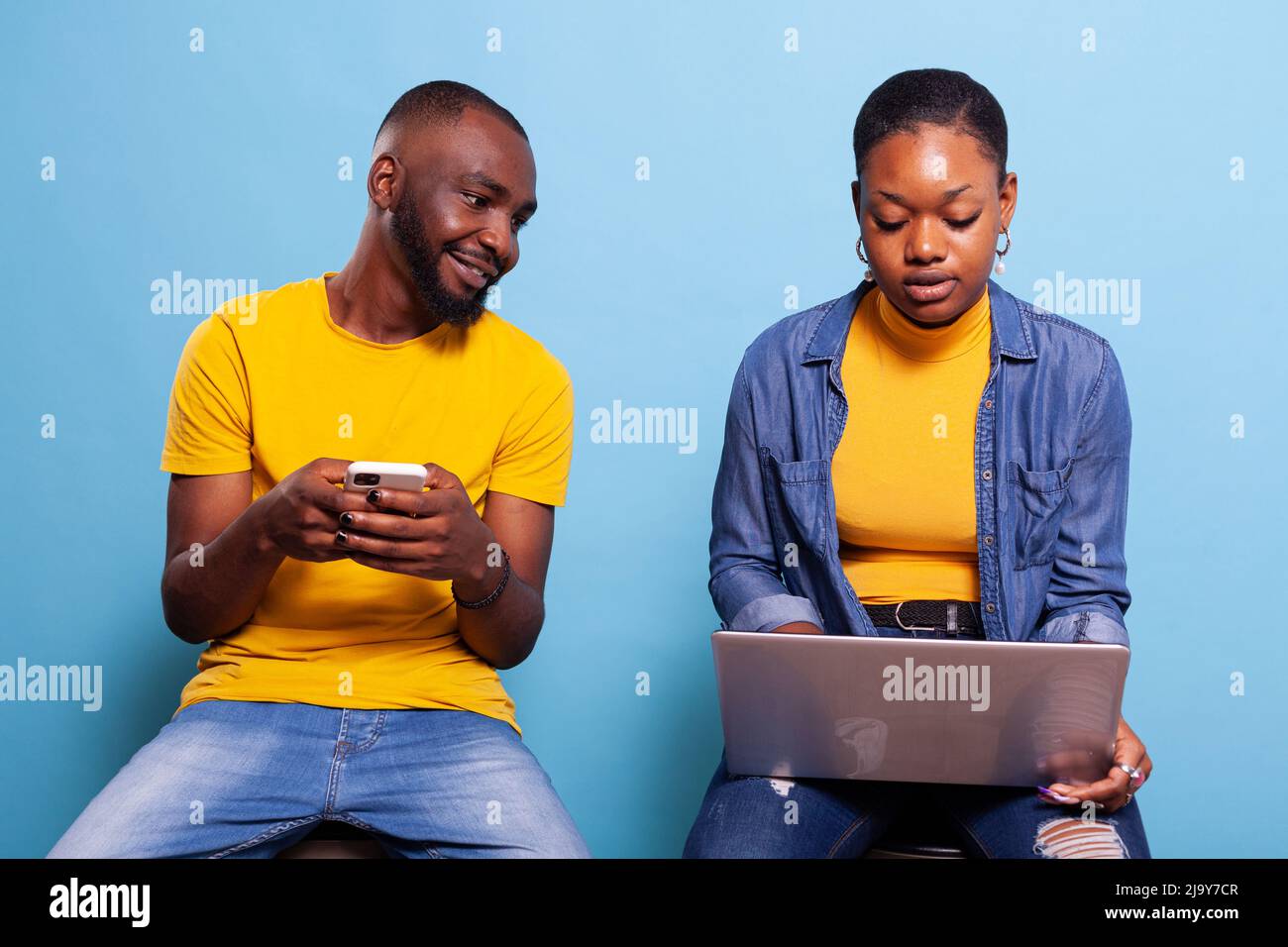 Les clients de la génération du millénaire utilisent un smartphone et un ordinateur portable en studio et s'amusent avec la technologie sur fond bleu. Homme et femme en relation regardant le téléphone mobile et l'ordinateur moderne. Banque D'Images