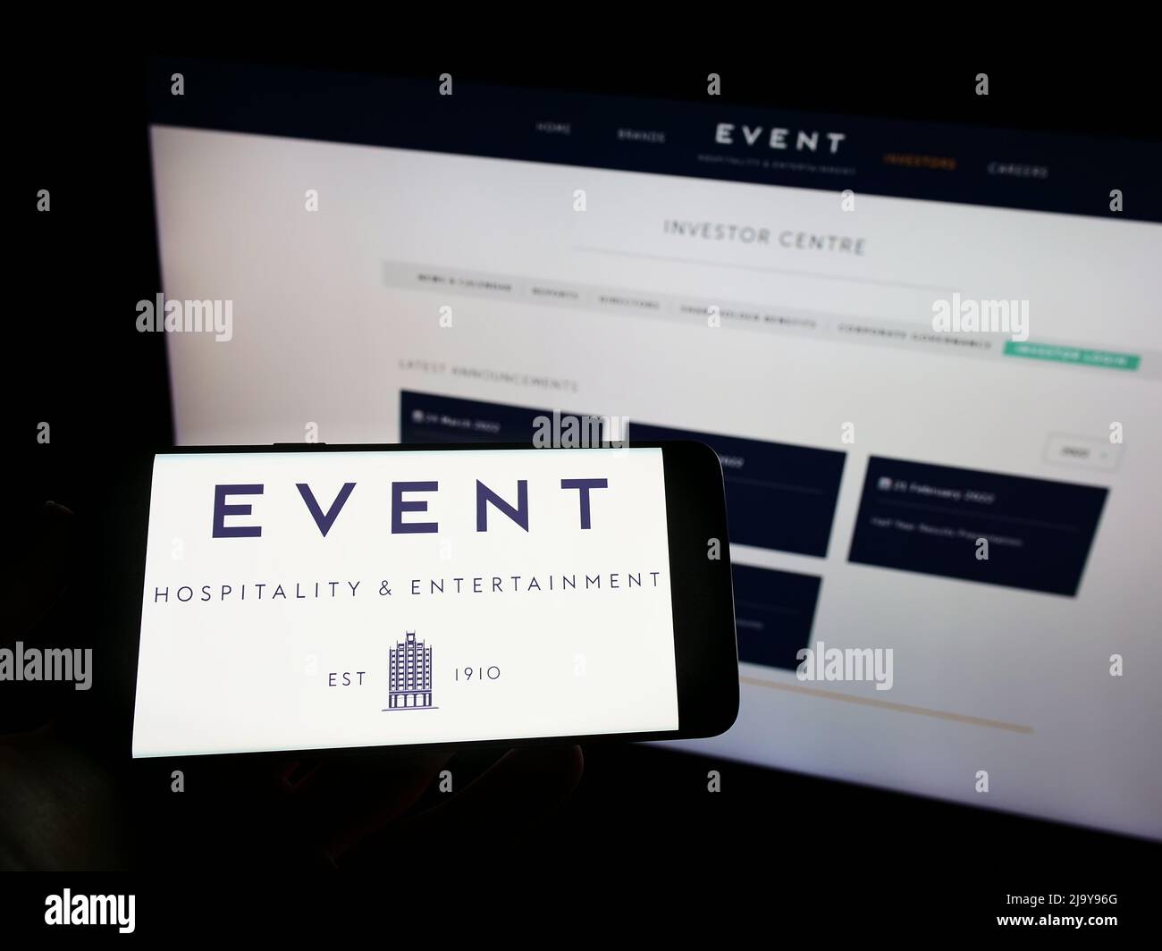 Personne tenant un téléphone portable avec le logo de la société Event Hospitality Entertainment Ltd. À l'écran en face de la page Web d'affaires. Mise au point sur l'affichage du téléphone. Banque D'Images