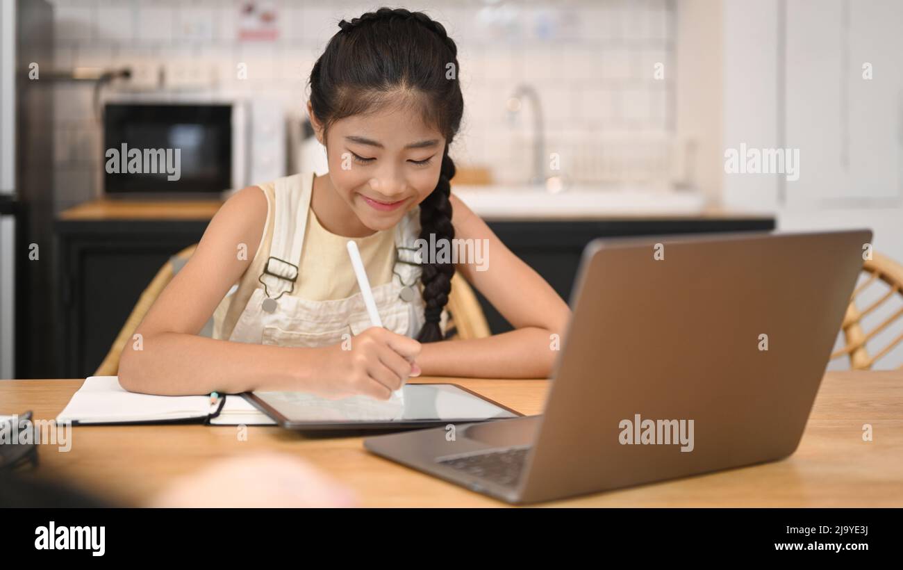 Adorable fille asiatique à distance apprendre en ligne, faisant des devoirs tout en étant assis à la table de cuisine Banque D'Images