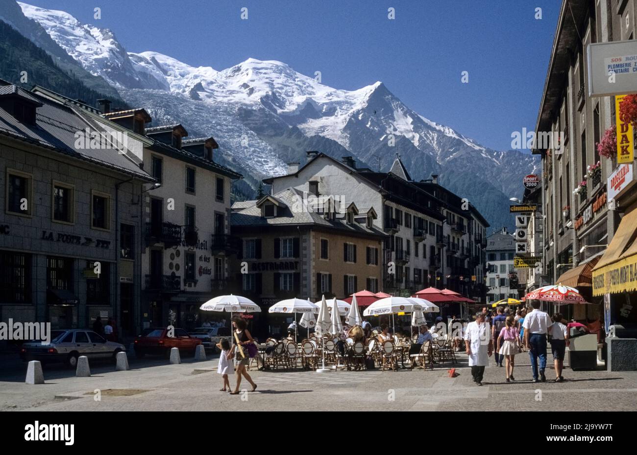 Cafés avec parasols, passants et voitures. Le massif du Mont blanc avec le Dôme du Gouter en arrière-plan. Place Balmat, Chamonix-Mont-blanc, France 1990 Banque D'Images