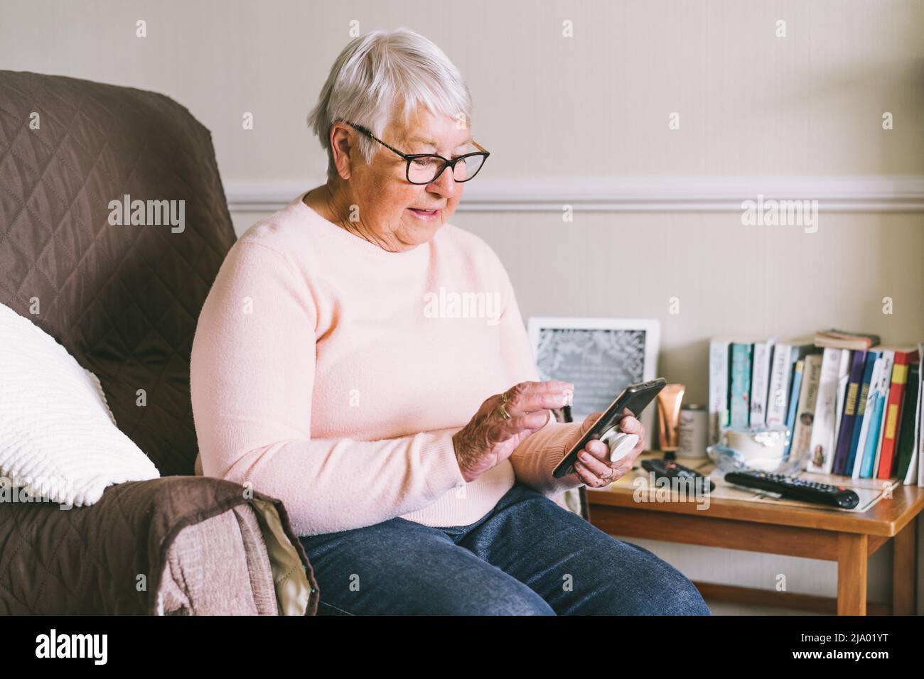 Femme âgée utilisant un smartphone assise sur un fauteuil à la maison. Une femme mûre et détendue tenant un téléphone portable SMS, acheter en ligne, jouer à des jeux, vérifier Banque D'Images Femme âgée utilisant un smartphone assise sur un fauteuil à la maison. Une femme mûre et détendue tenant un téléphone portable SMS, acheter en ligne, jouer à des jeux, vérifier Banque D'Images
