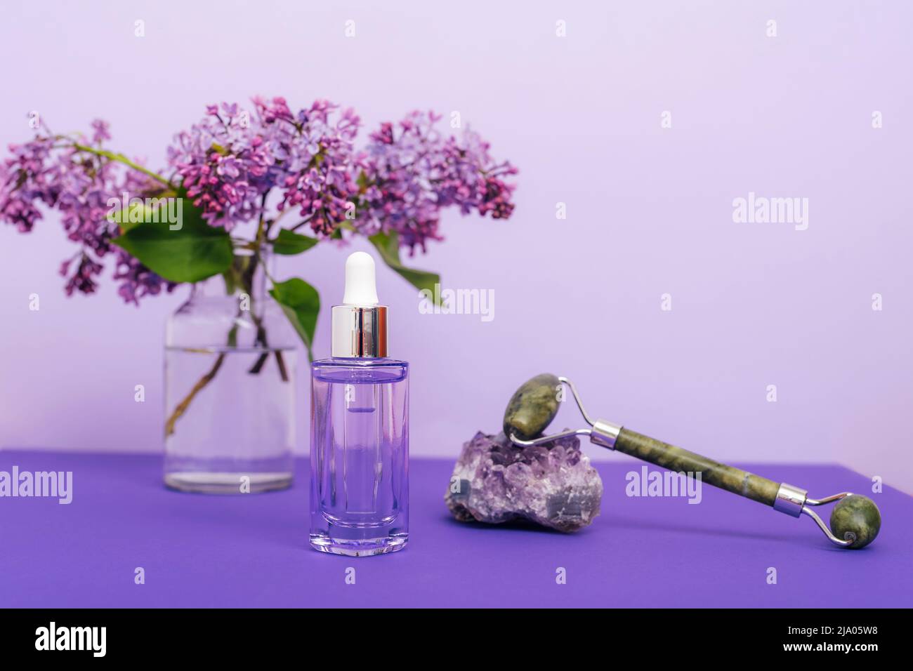 Flacon de sérum cosmétique, rouleau facial jade et fleurs lilas sur fond violet. Cosmétiques naturels, concept de soins de la peau. Banque D'Images