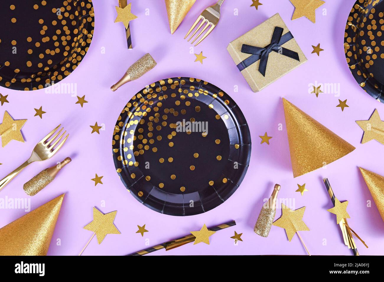 Plat de fête avec assiettes noires et dorées, fourchettes, cadeau, bouteille de champagne et confetti aux étoiles sur fond violet Banque D'Images