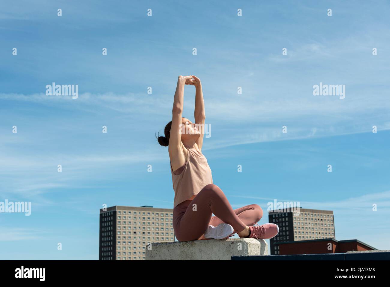 Femme sportive assise et s'étirant avec ses bras au-dessus de sa tête, en milieu urbain. Banque D'Images