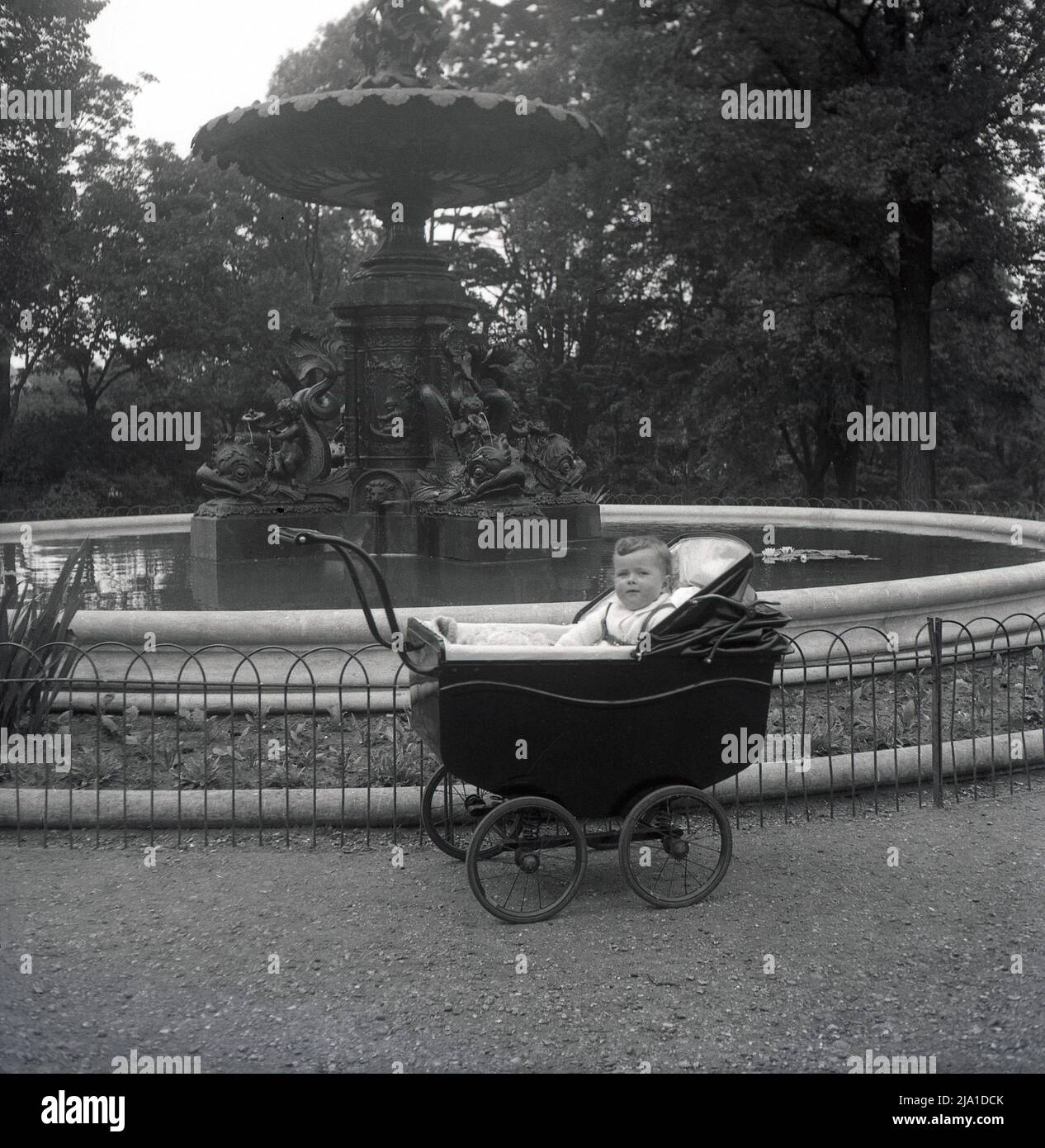 1950s, historique, un enfant en bas âge assis dans un petit pram de l'époque construit par un autocar à roues, à côté de la fontaine John Woodward Memorial à Dane Park, Margate, Kent, Angleterre, Royaume-Uni. La fontaine en fonte a été érigée en 1896 à la mémoire du don de la terre sur laquelle le parc a été créé. Banque D'Images