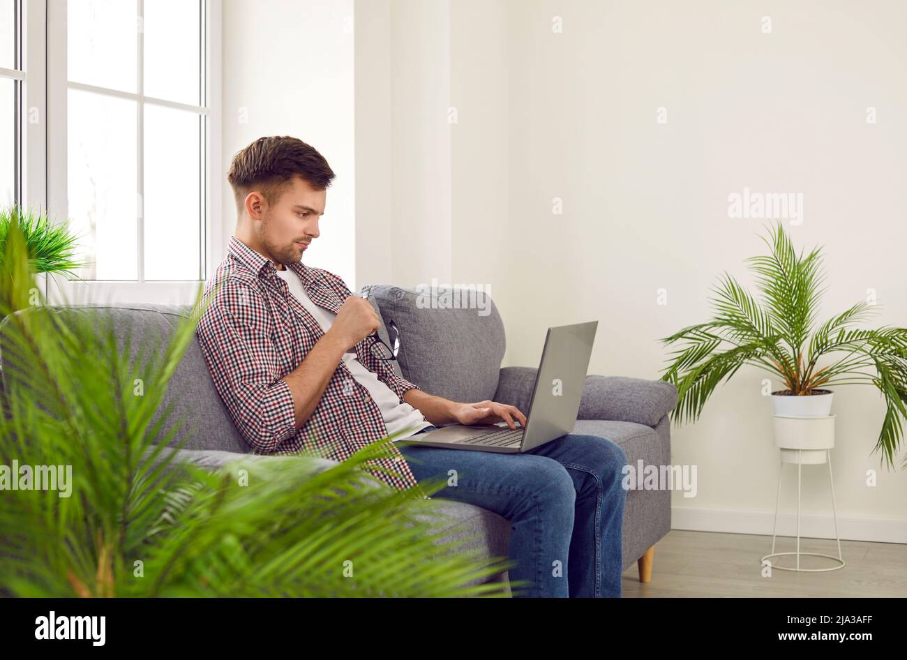 Jeune homme indépendant avec une expression faciale sérieuse travaillant à distance à la maison à l'aide d'un ordinateur portable. Banque D'Images