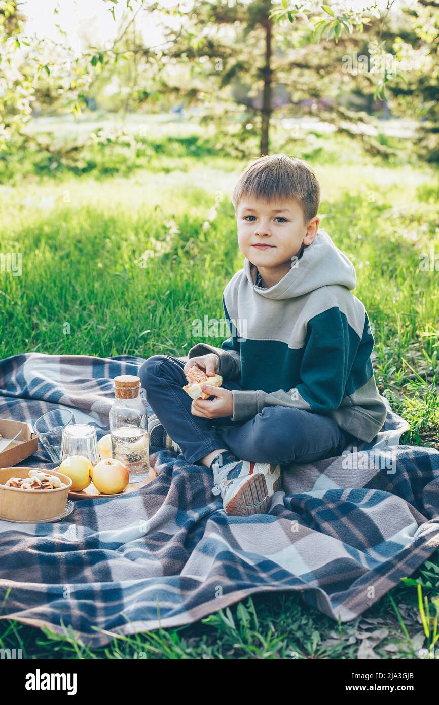 Garçon de 7-8 ans assis sur un plat et mangeant de la pizza. Concept de pique-nique de printemps ou d'été Banque D'Images