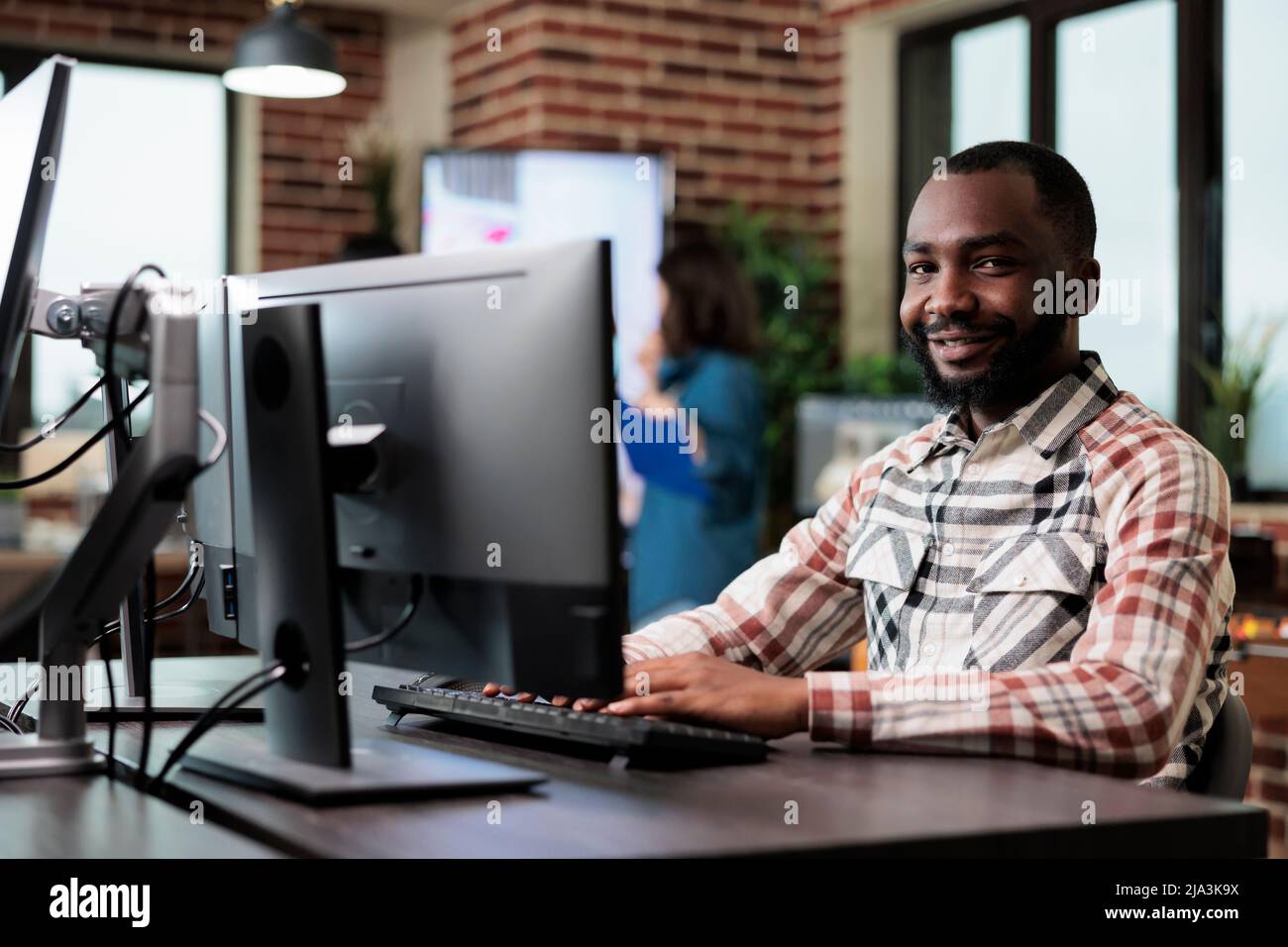 Agence adulte souriant travailleur heureux à l'ordinateur dans le studio de démarrage espace intérieur moderne. Un employé d'une entreprise créative afro-américaine assis à un bureau dans un espace de travail tout en regardant la caméra. Banque D'Images