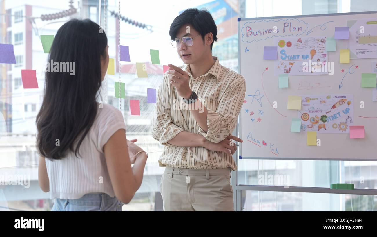 Jeune femme créative se tenant près du tableau de conférence avec son collègue et discutant ensemble de la nouvelle stratégie de marketing. Banque D'Images