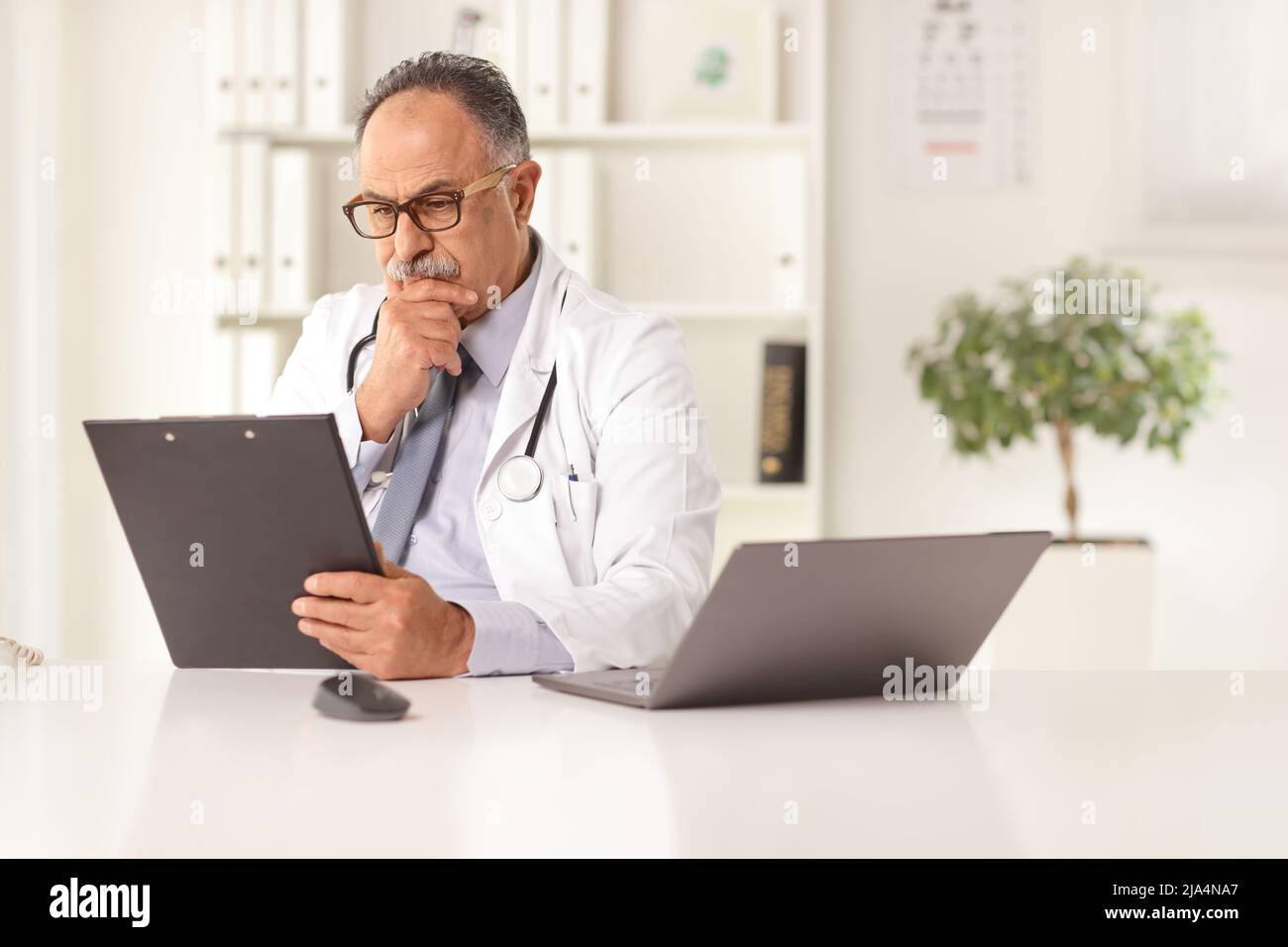 Un homme mature médecin assis dans un bureau et lisant un document sur un bureau avec un ordinateur portable Banque D'Images