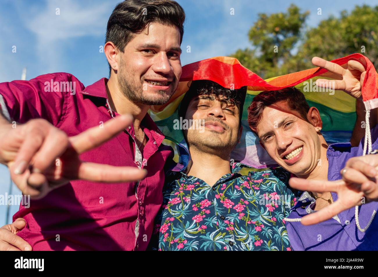 portrait latino gay hommes argentins amis, avec le drapeau de fierté gay sur leur tête faisant le signe v avec les mains, en plein air souriant et regardant l'appareil photo Banque D'Images