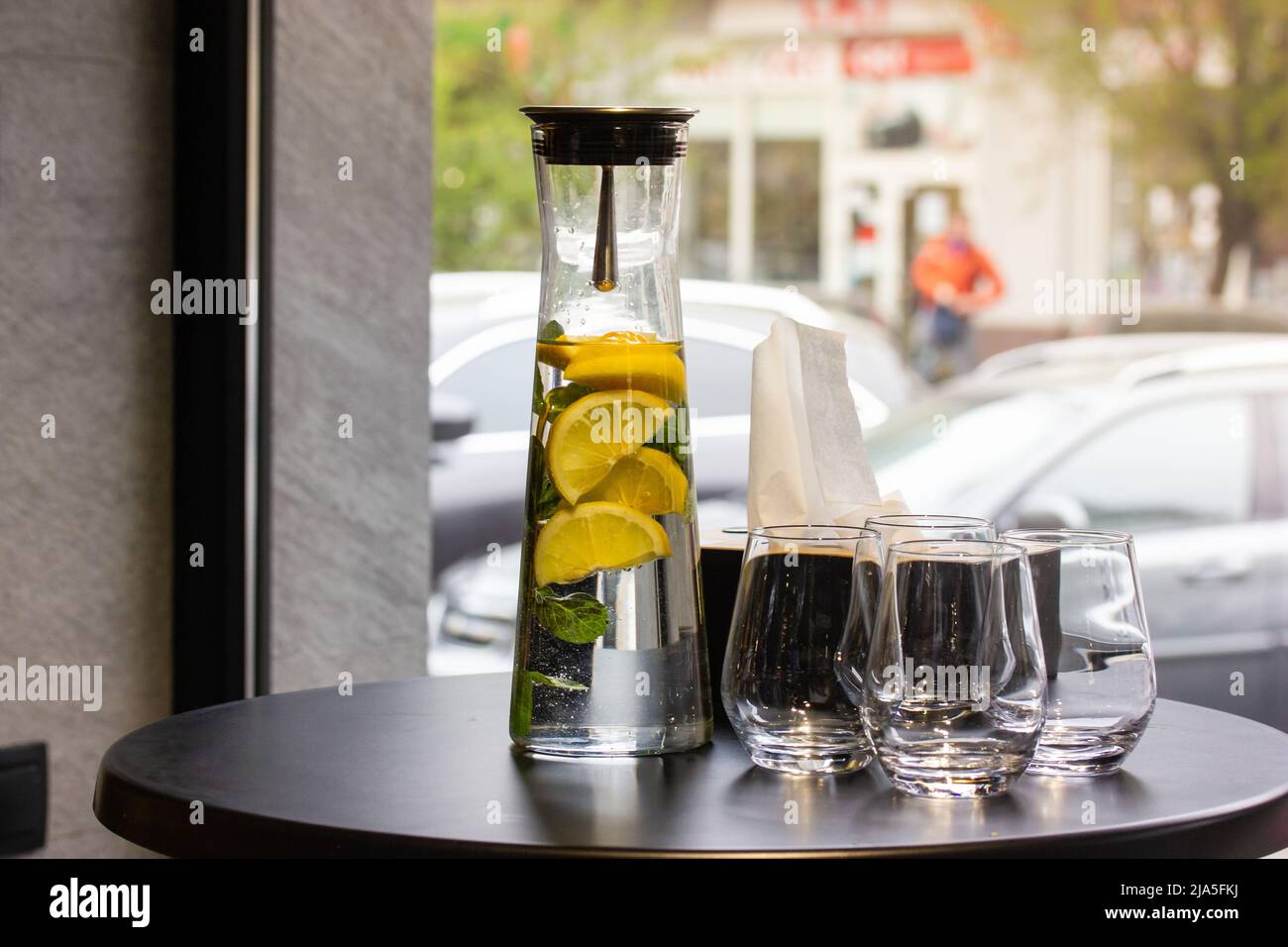 Une carafe en verre transparent, un pichet de limonade avec des citrons, de la menthe à côté des verres sur une table ronde noire dans un café ou un restaurant contre une fenêtre panoramique. A r Banque D'Images