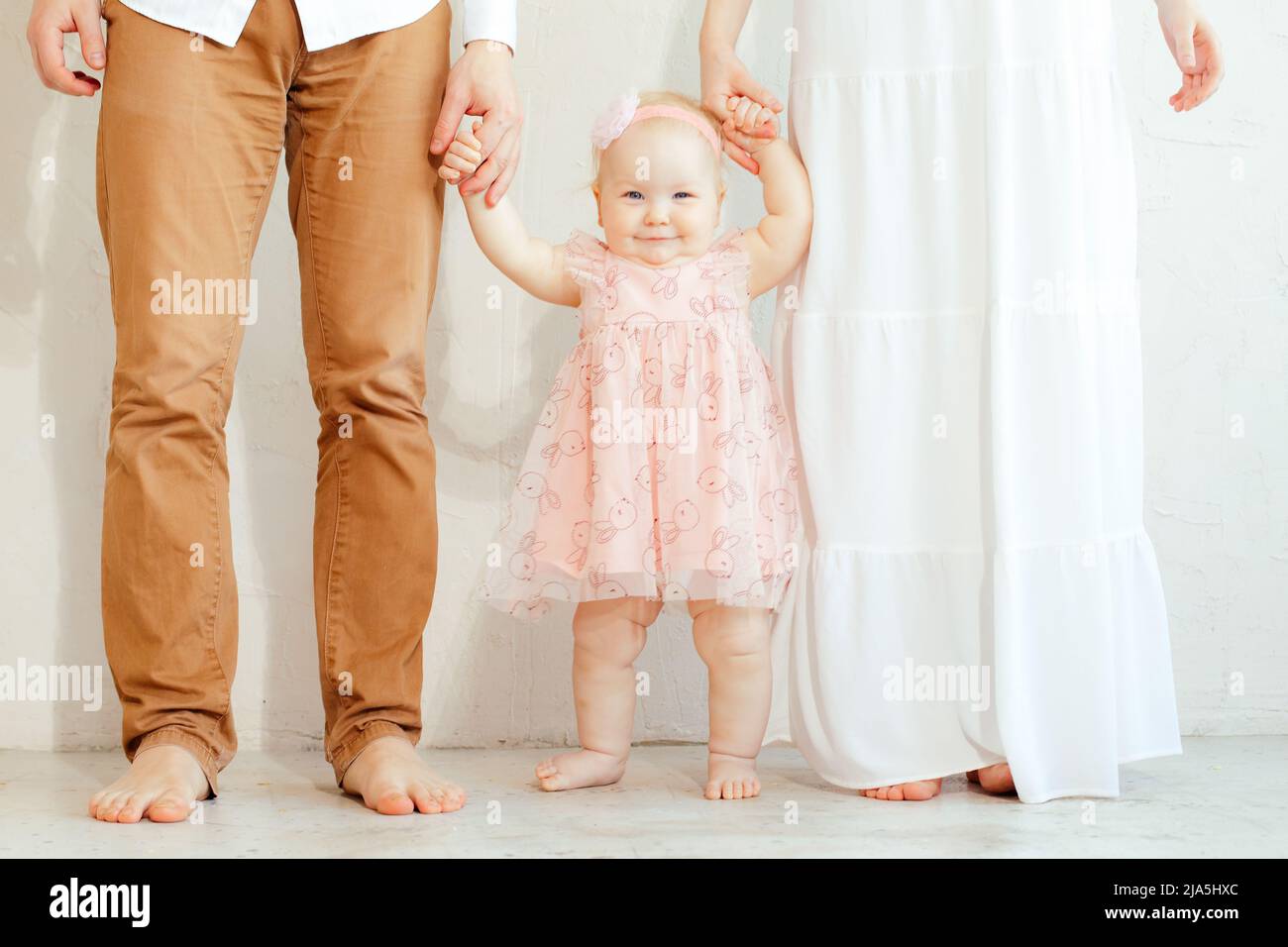 Portrait de mère et de père sans chaussures tenant les mains de la jeune fille souriante en robe de vacances rose sur fond blanc. Premier anniversaire de fête, heureux Banque D'Images