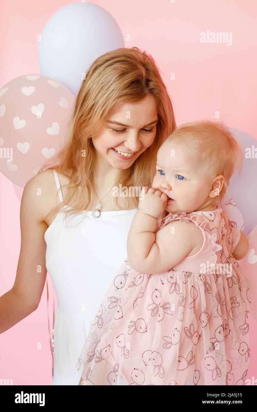 Portrait gros plan de la jeune mère souriante tenant une petite fille mignonne dans la robe de vacances sur fond rose. Première fête d'anniversaire, décor rose et ballons Banque D'Images