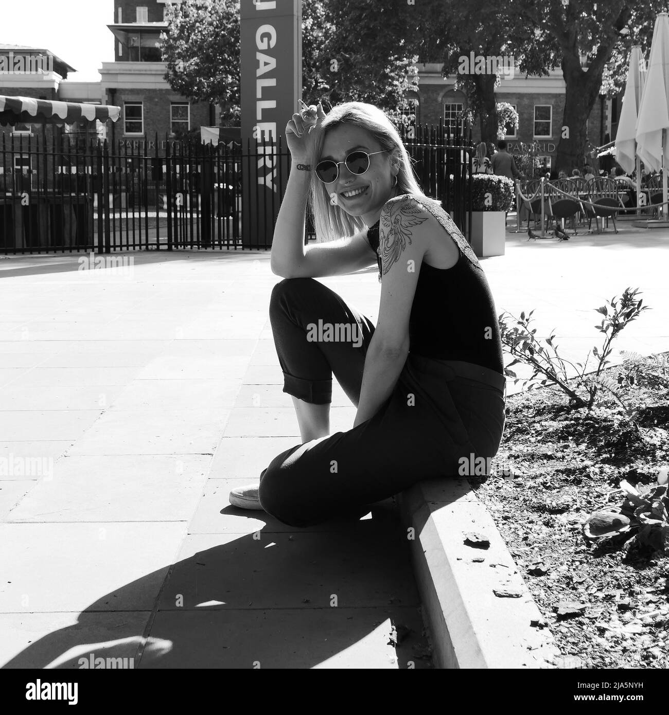 Jolie dame avec des tatouages, le perçage du nez et des lunettes de soleil se trouve sur une pierre de trottoir, sourit et fume. Duke of York Square, Chelsea, Londres. Monochrome. Banque D'Images