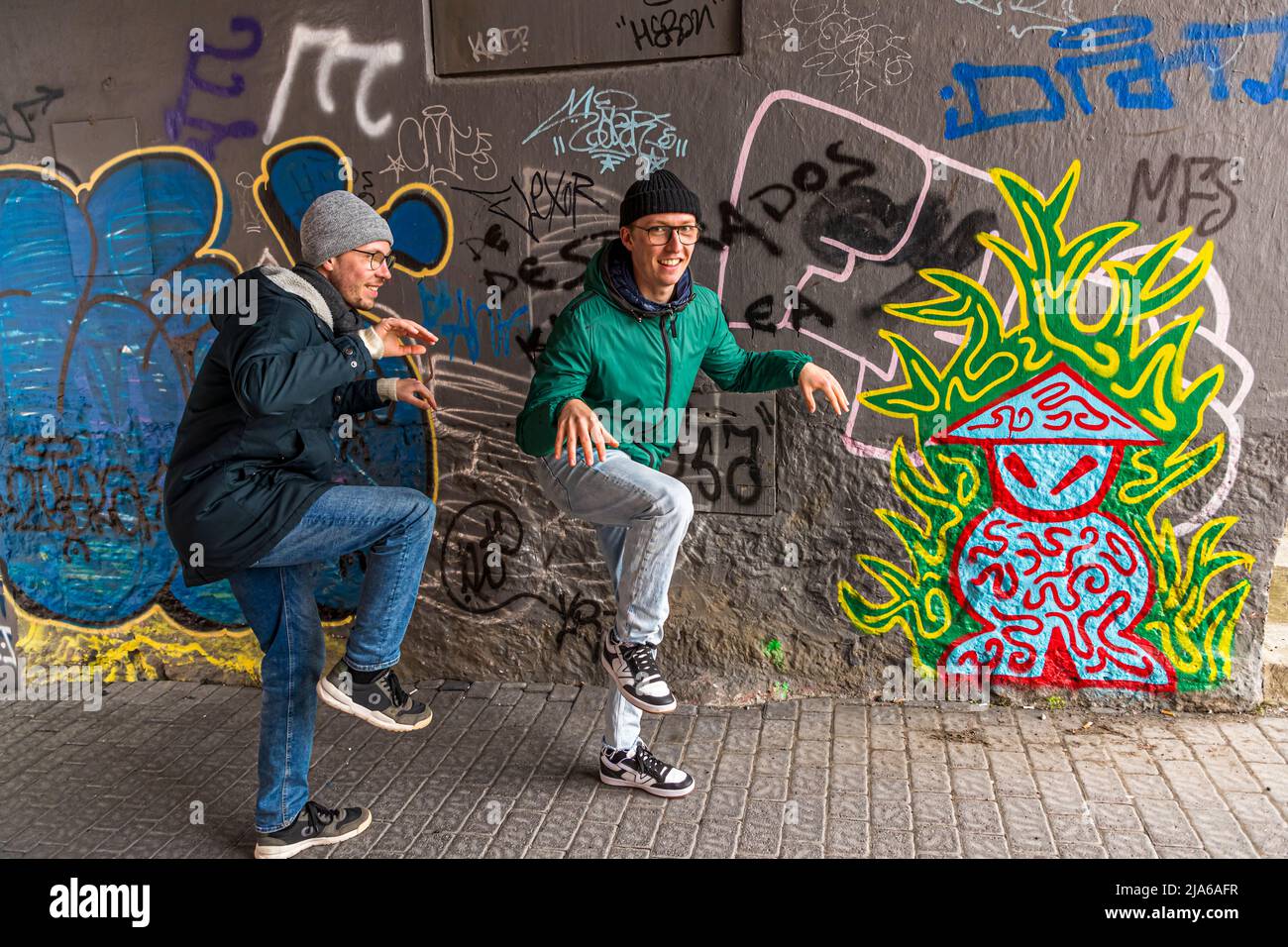 Deux jeunes hommes posent ici de manière ludique avec une jambe levée devant une fresque colorée représentant une figure fantastique stylisée sur un mur de maison à Reykjavik. Banque D'Images