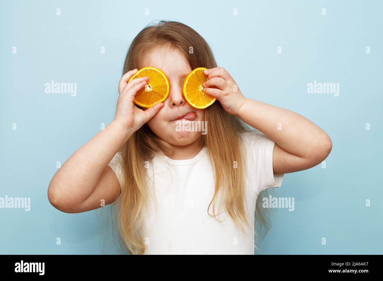 petite fille heureuse avec des moitiés d'oranges couvrant les yeux sur un fond bleu. enfant amusant et montrant la langue. Banque D'Images